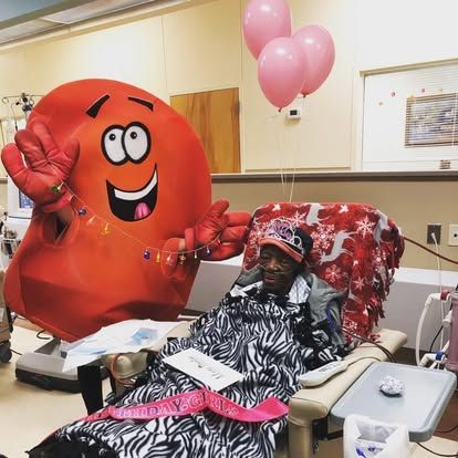 Smiling red balloon character beside a hospitalized patient in a decorated room with pink balloons