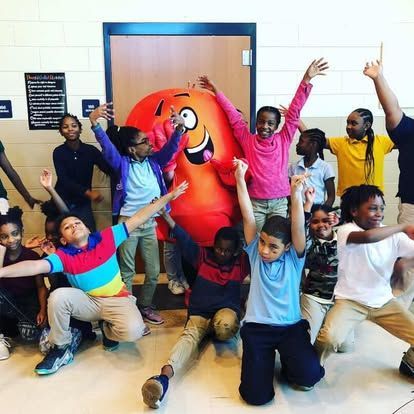 Children posing with a large orange mascot in a hallway, smiling and raising their arms