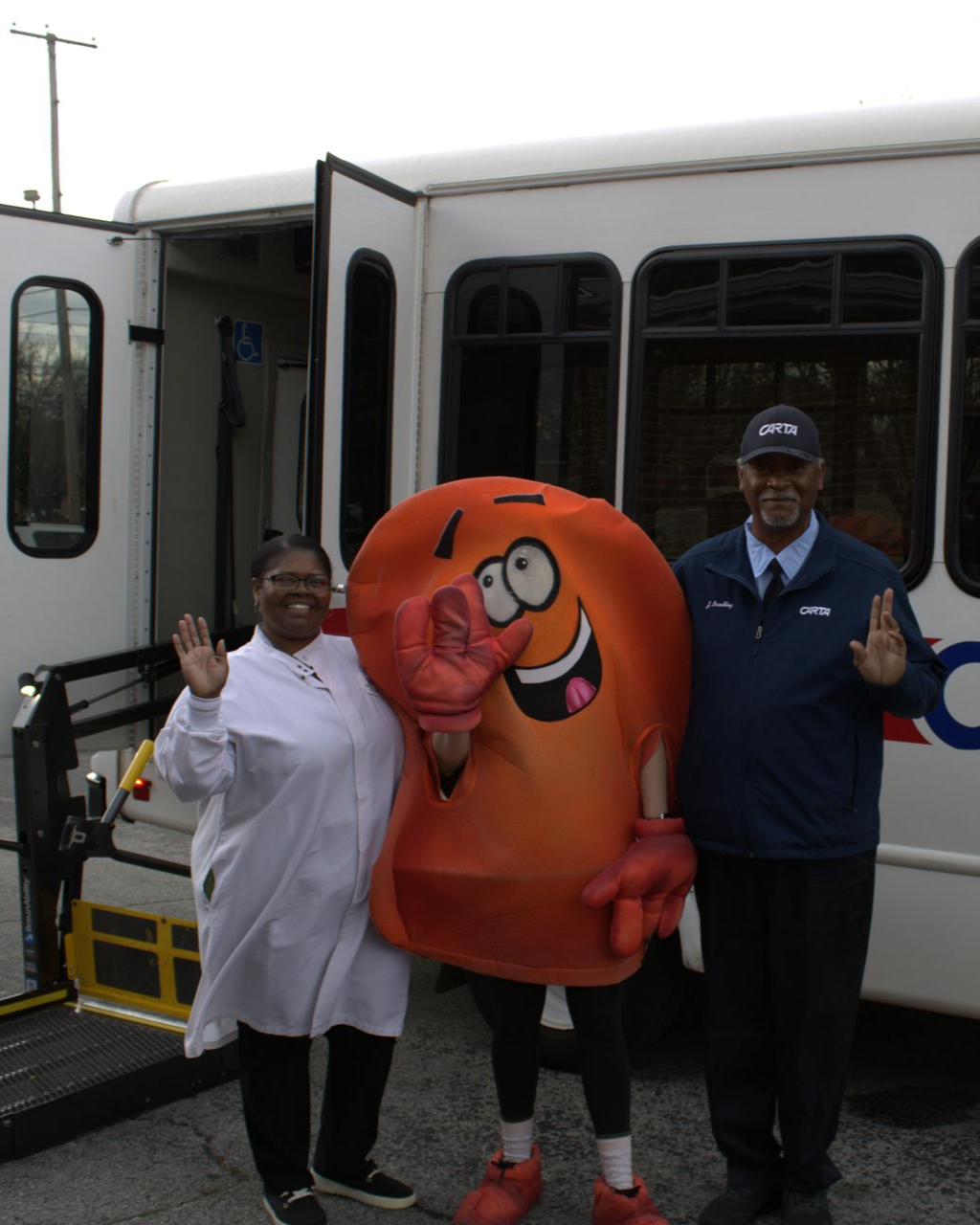 Three people pose beside a white bus, including an orange mascot giving thumbs up.