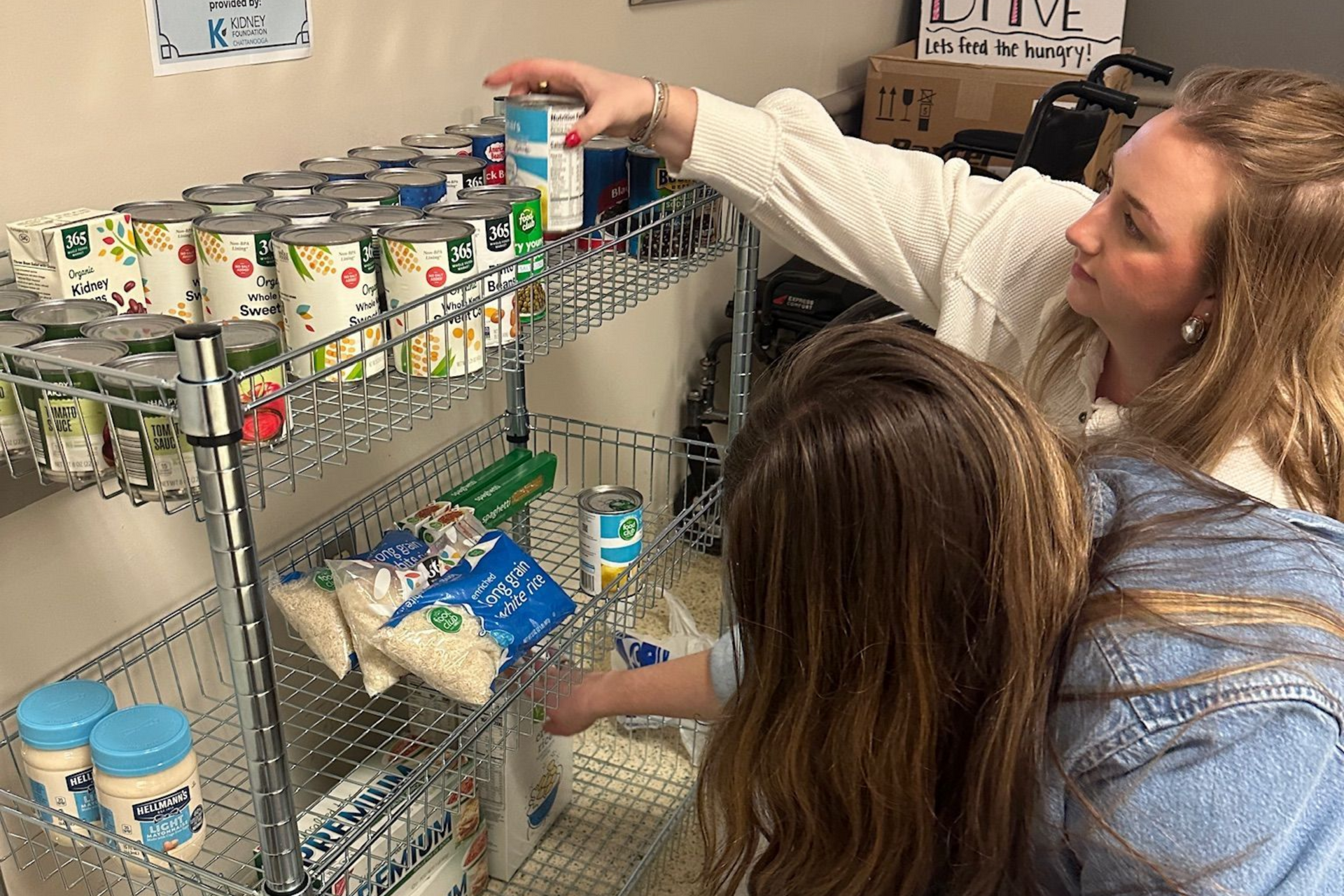 Two people stock a pantry shelf with canned goods and packaged food.