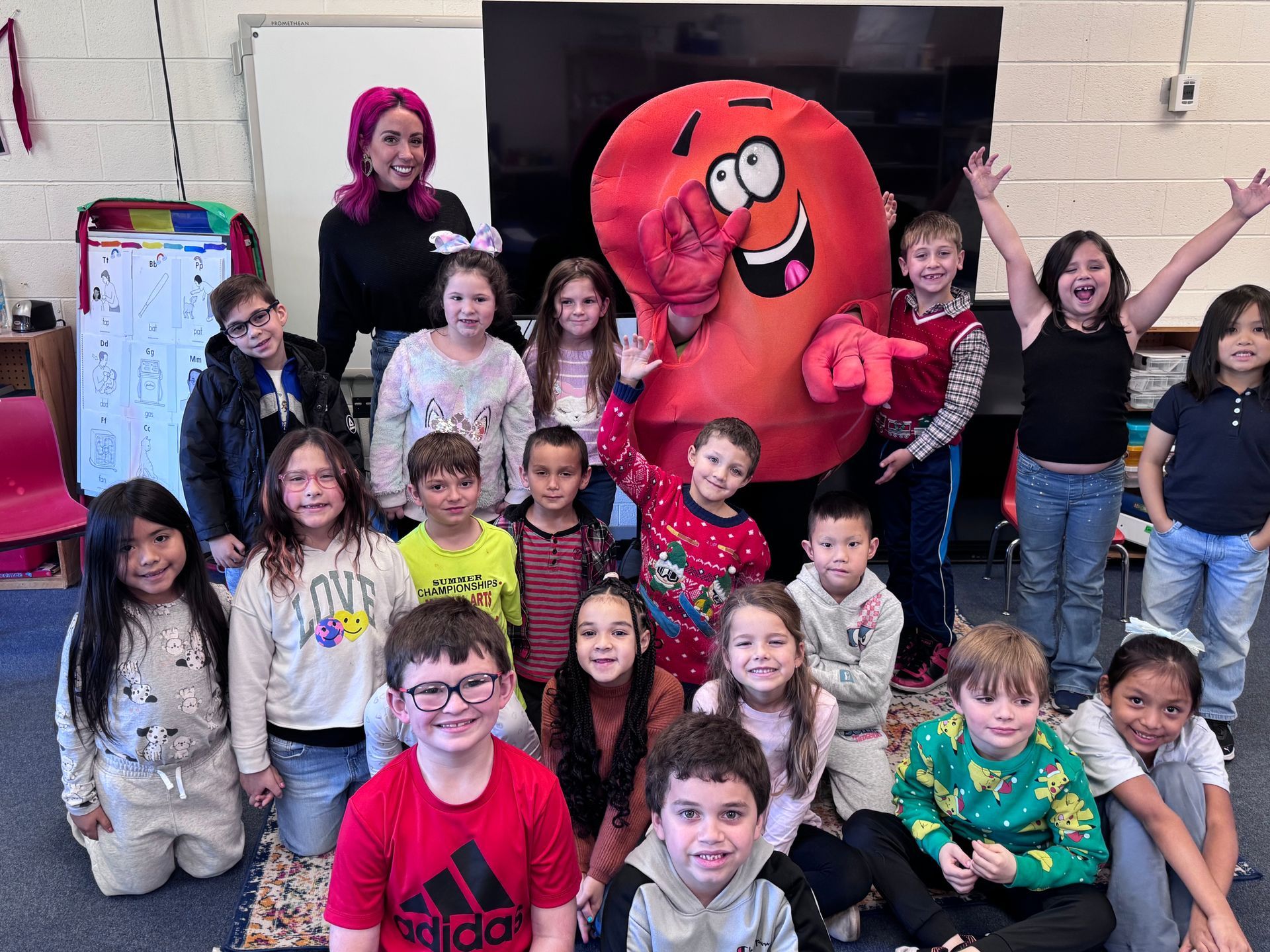 Group of children posing with a large red mascot in a classroom, some smiling and waving.