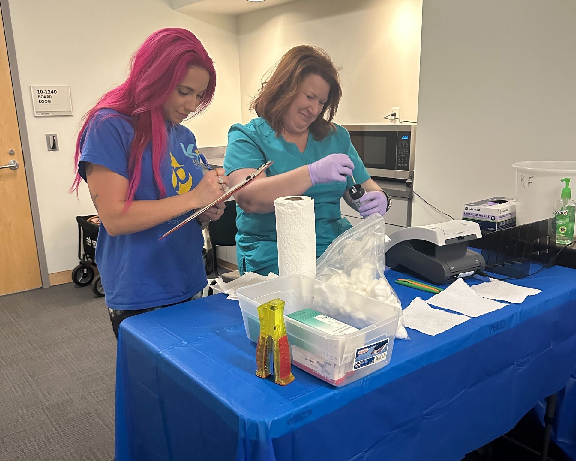 Two people in blue shirts preparing materials at a table with gloves, bags, and containers in a room.