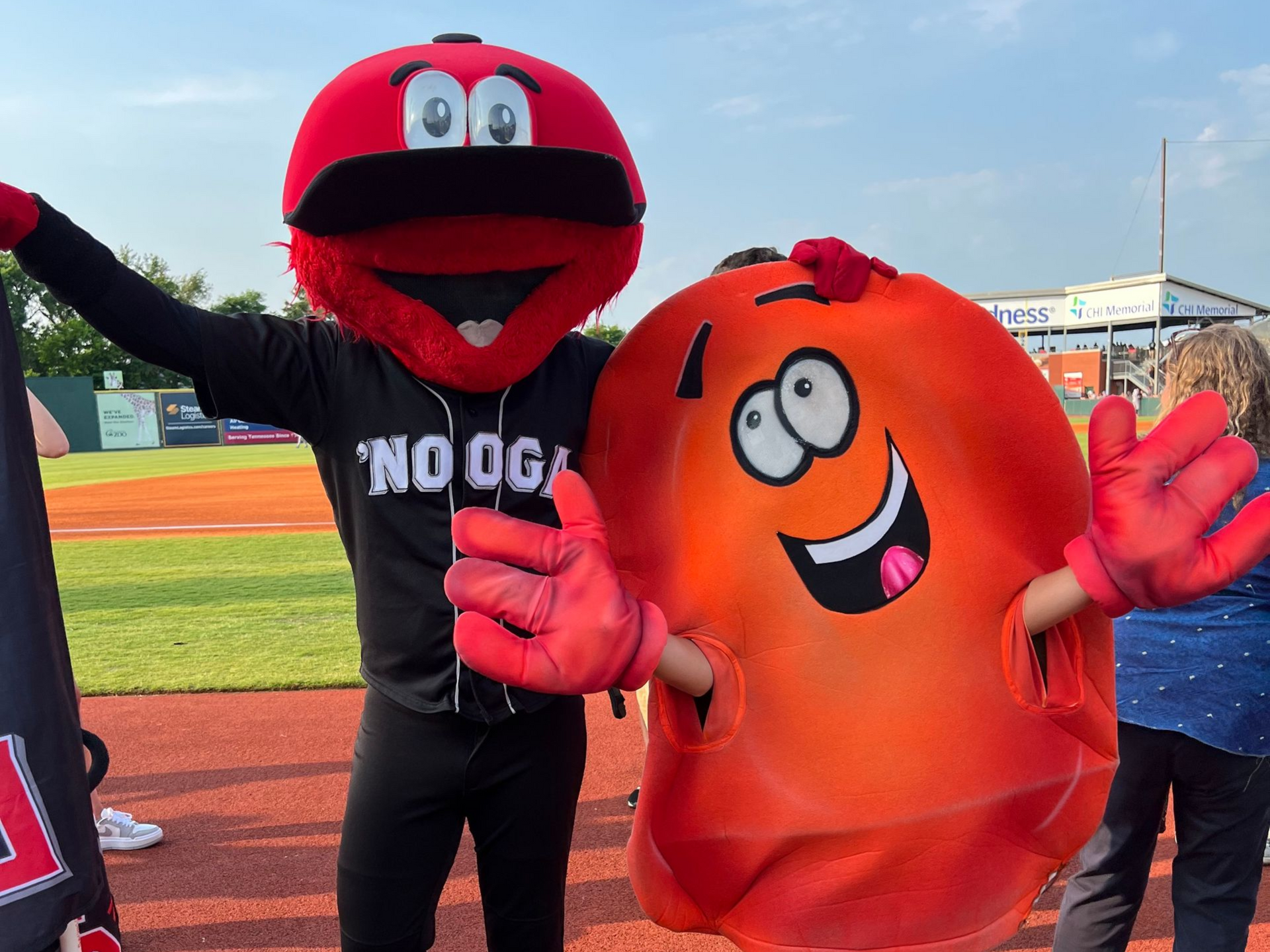 Two colorful mascots posing on a sports field, one red and one orange, both waving.