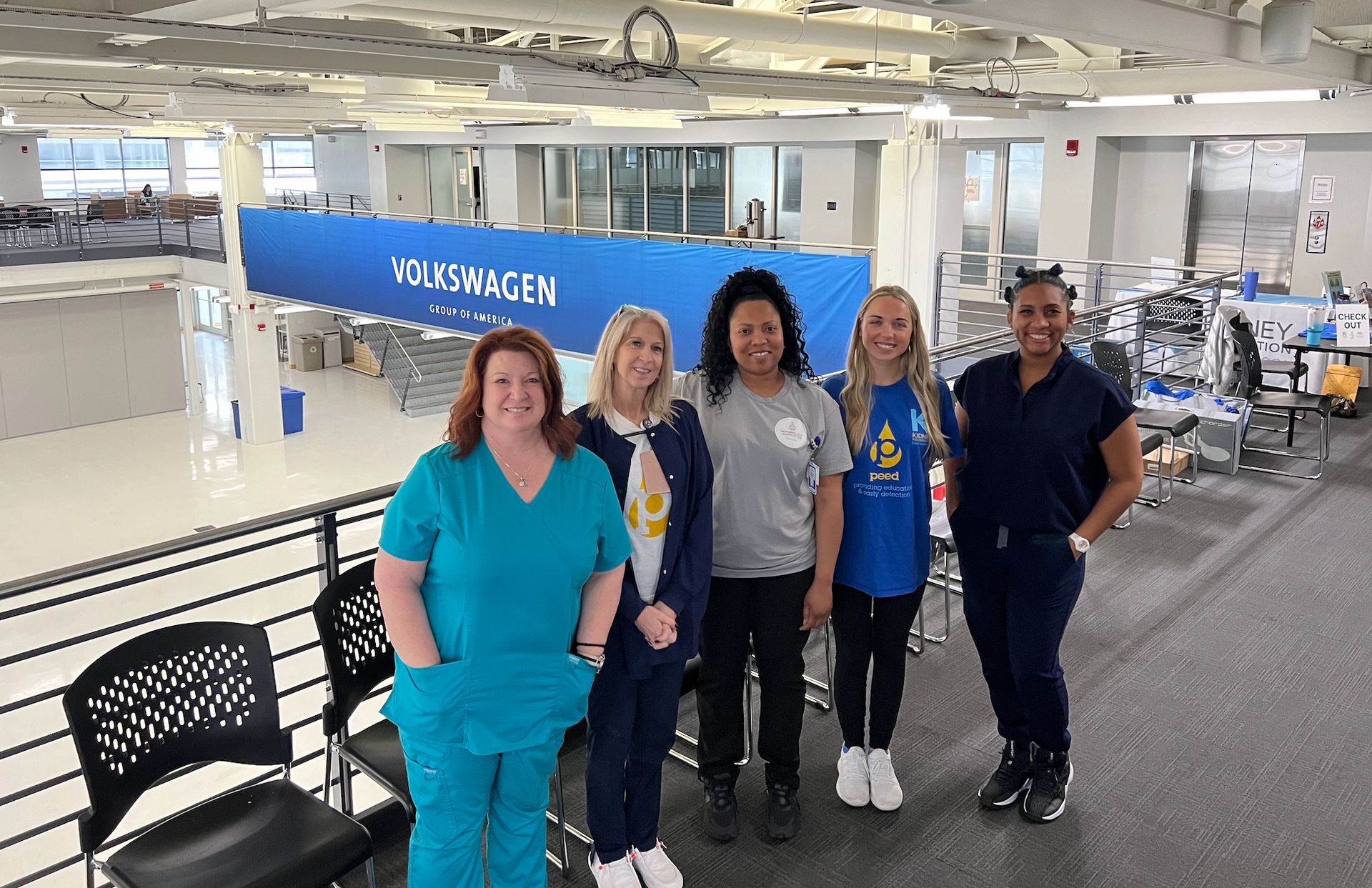 Five people posing indoors beside a blue banner in a bright open office space