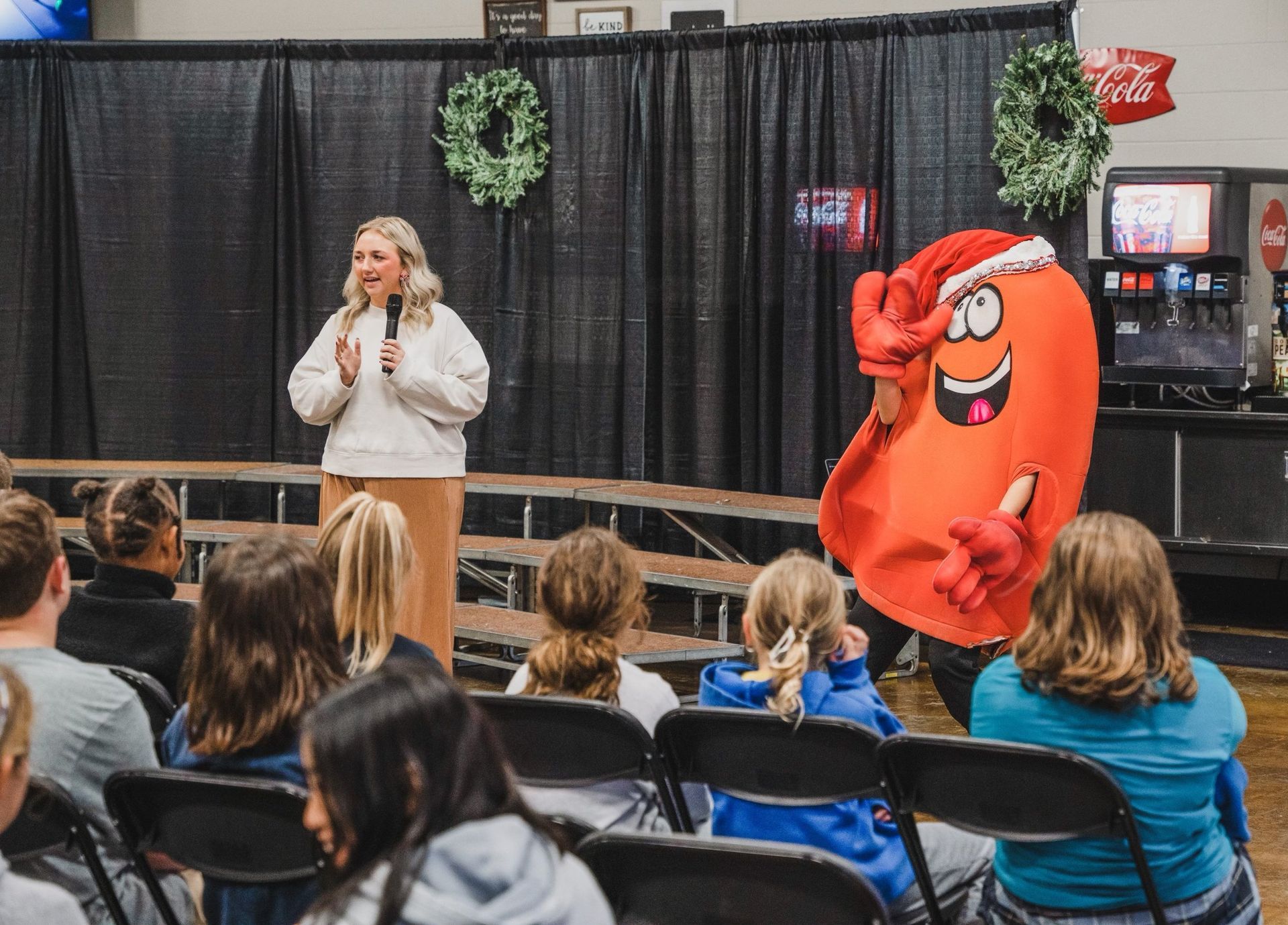 Woman speaks on stage beside red mascot costume, facing an audience in a hall.