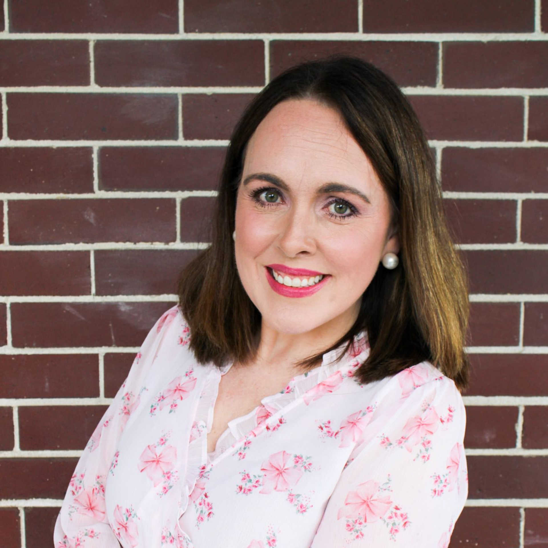 Smiling woman with shoulder-length dark hair in a white floral blouse against a brick wall