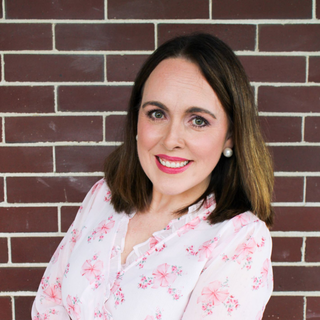 Smiling woman with shoulder-length dark hair in a white floral blouse against a brick wall