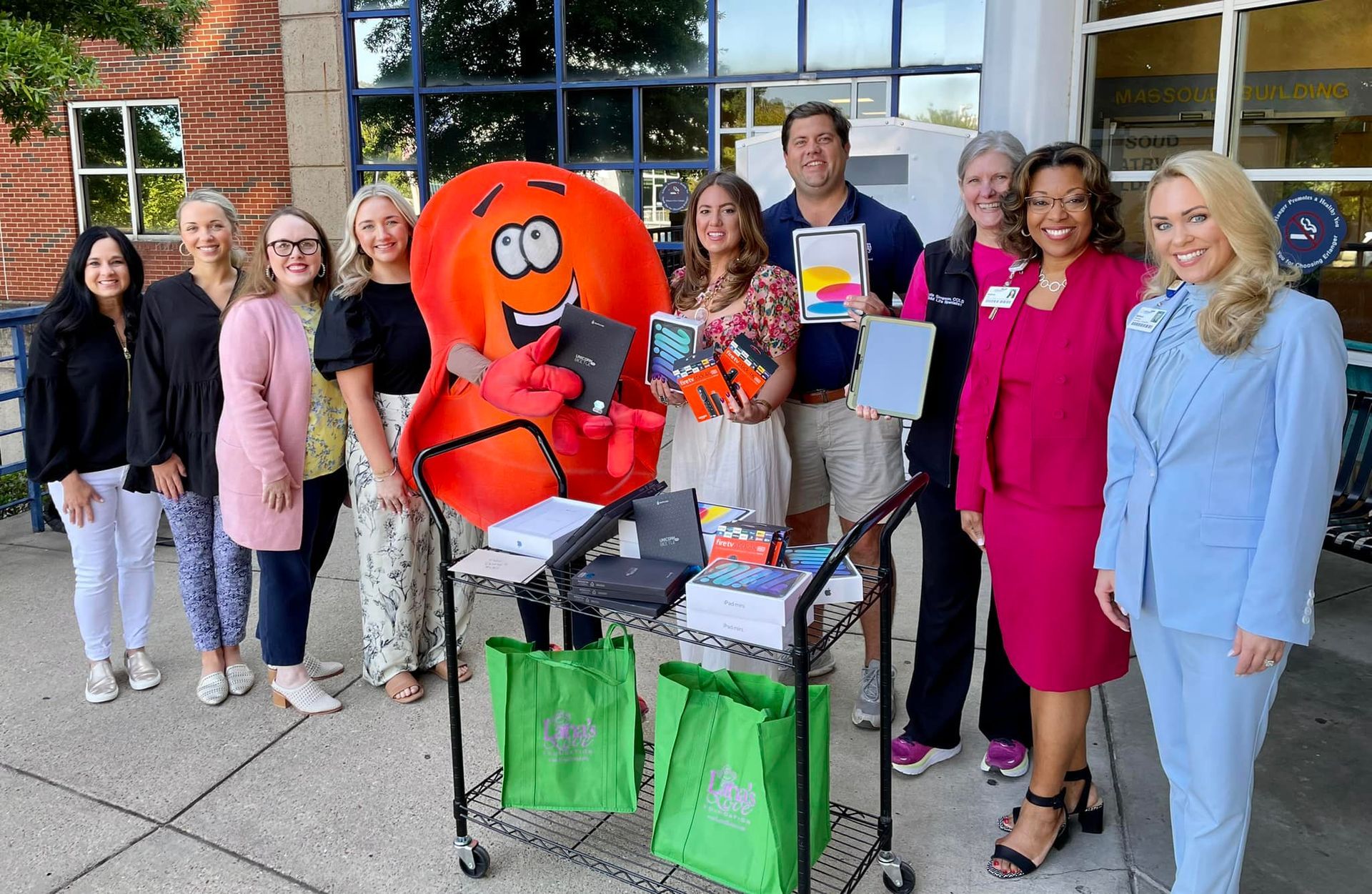 Group posing with an orange mascot beside a cart of green bags outside a building