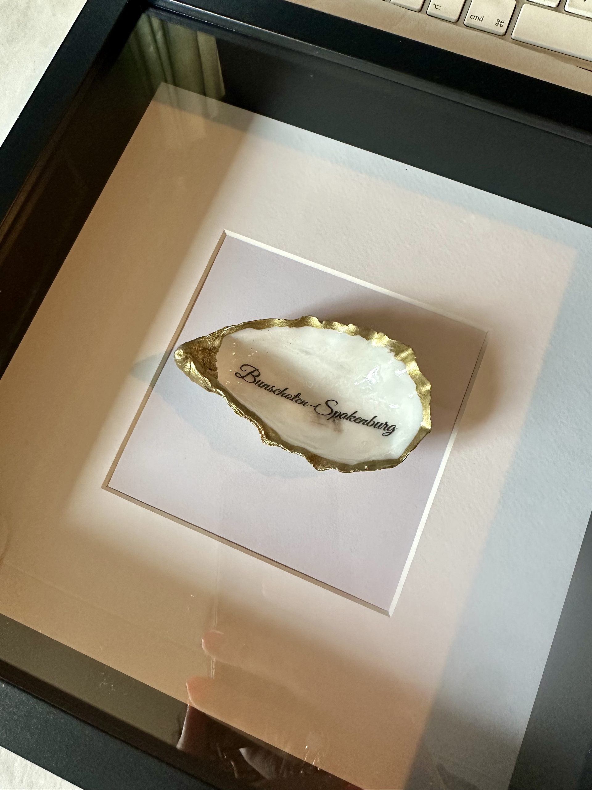 Een decoratieve oesterschelp met goudkleurige randen en sierlijk schrift, ingelijst in een klein zwart lijstje.