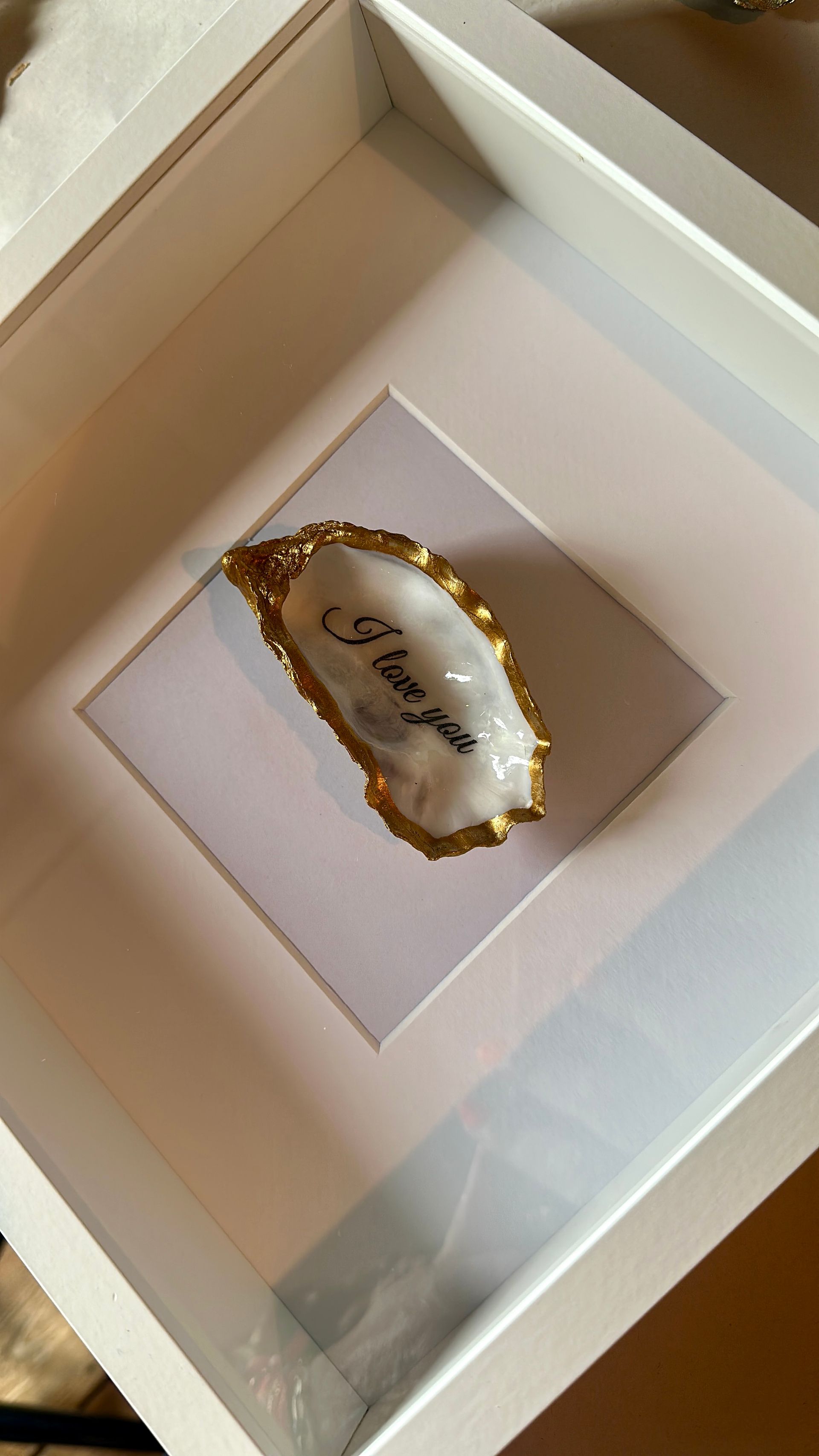 Een ingelijste vitrine met een witte oesterschelp met goudkleurige randen en kalligrafie, gecentreerd op een witte achtergrond.