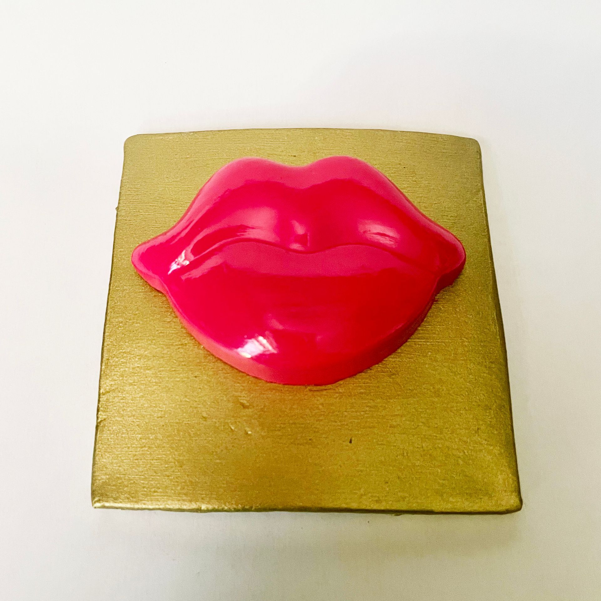 Een roze, lippenvormig object op een goudkleurig vierkant oppervlak.