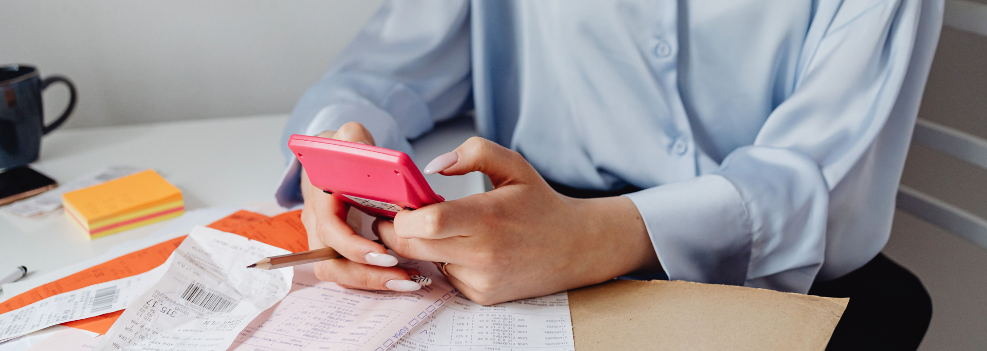 Picture of hands holding a pink smartphone at a desk with papers, notebooks, and an orange pen.