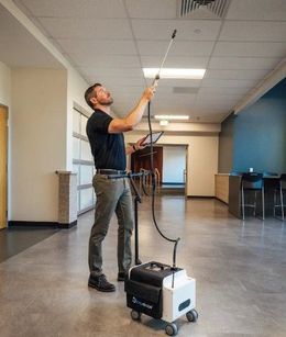 Man adjusting a ceiling fixture with a long tool in a hallway, standing on a floor machine with cables attached