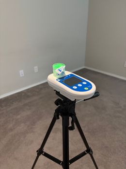 Tripod-mounted device with a small green container in an empty room with beige walls and carpet.