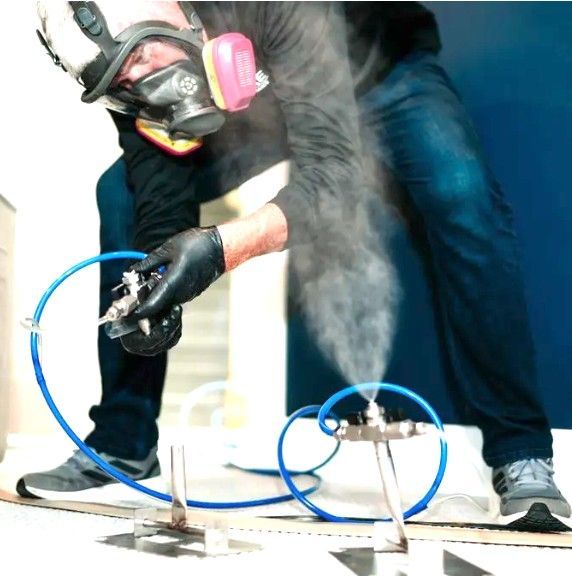 Person in a gas mask sprays a surface with a hose near blue tubing and metal stands