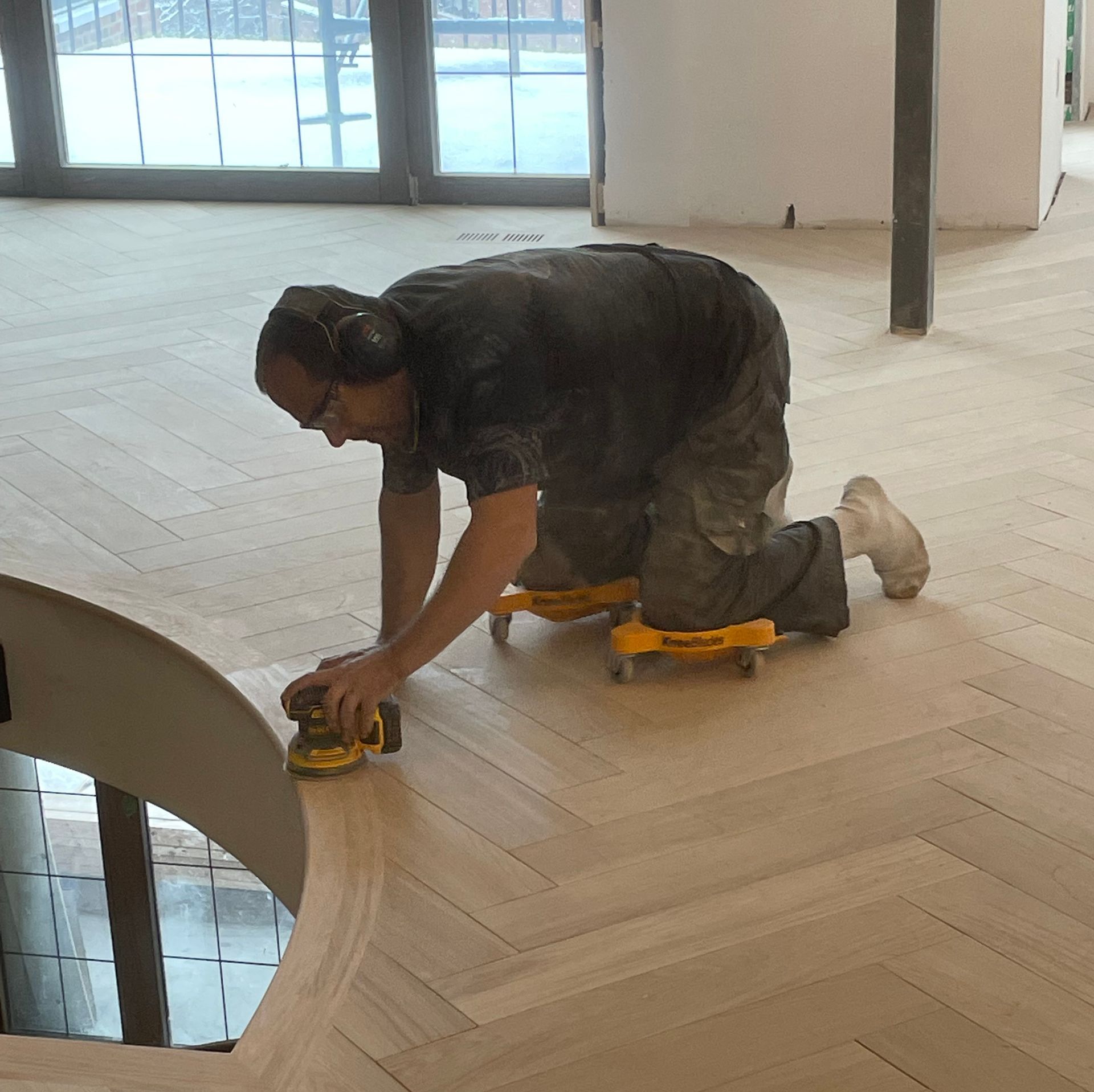 Un homme agenouillé utilise une ponceuse électrique sur un sol en bois dans un espace intérieur avec de grandes fenêtres.