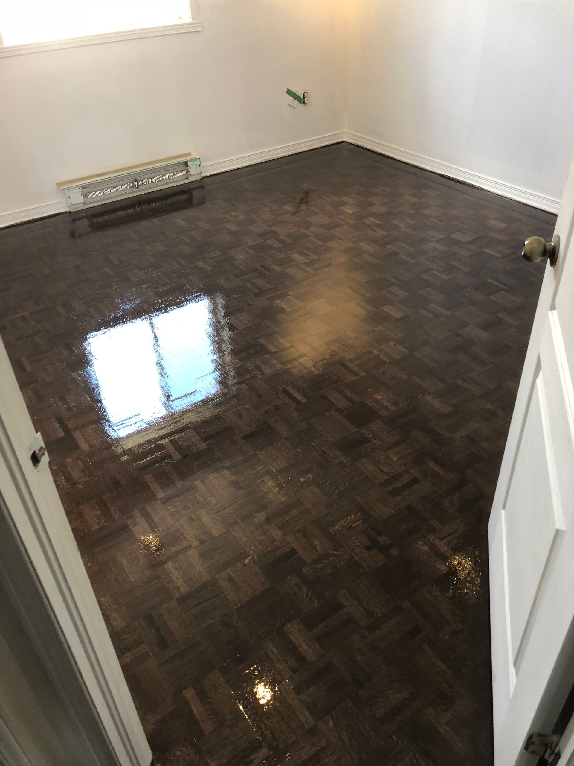 Une pièce vide avec des murs blancs, un sol en parquet brun foncé brillant, une fenêtre et des outils de construction.