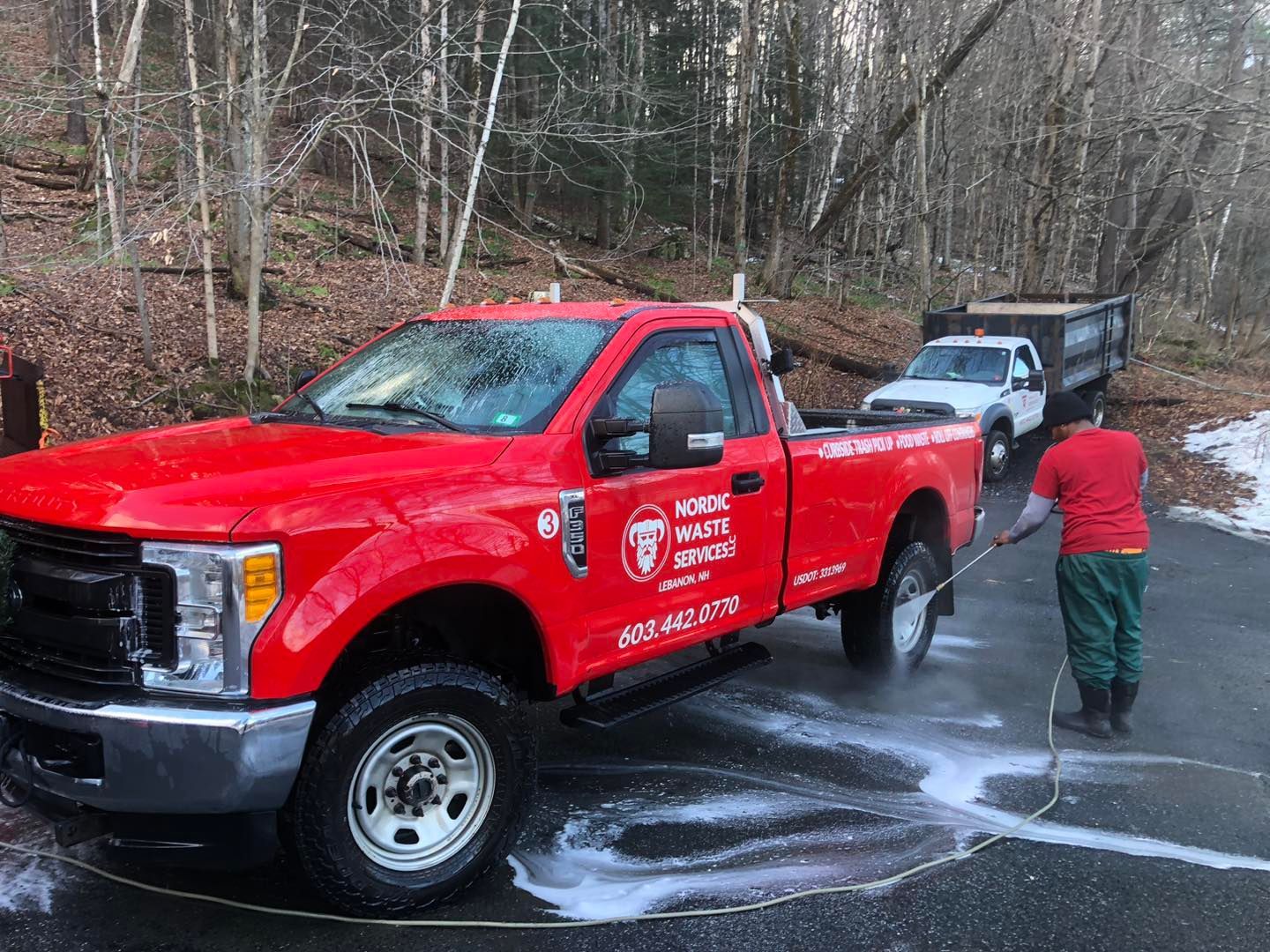 Red truck being washed by a person on a paved surface near two other trucks in a wooded area.