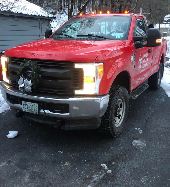 Red Nordic Waste Services truck with a wreath on the front, parked on a snowy driveway.