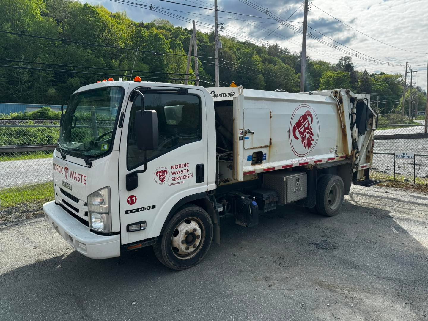 White garbage truck with open door, parked on asphalt, tree-lined background.