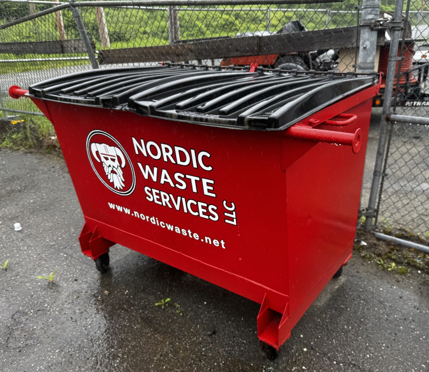 Red Nordic Waste Services dumpster with black lid, near a chain-link fence.