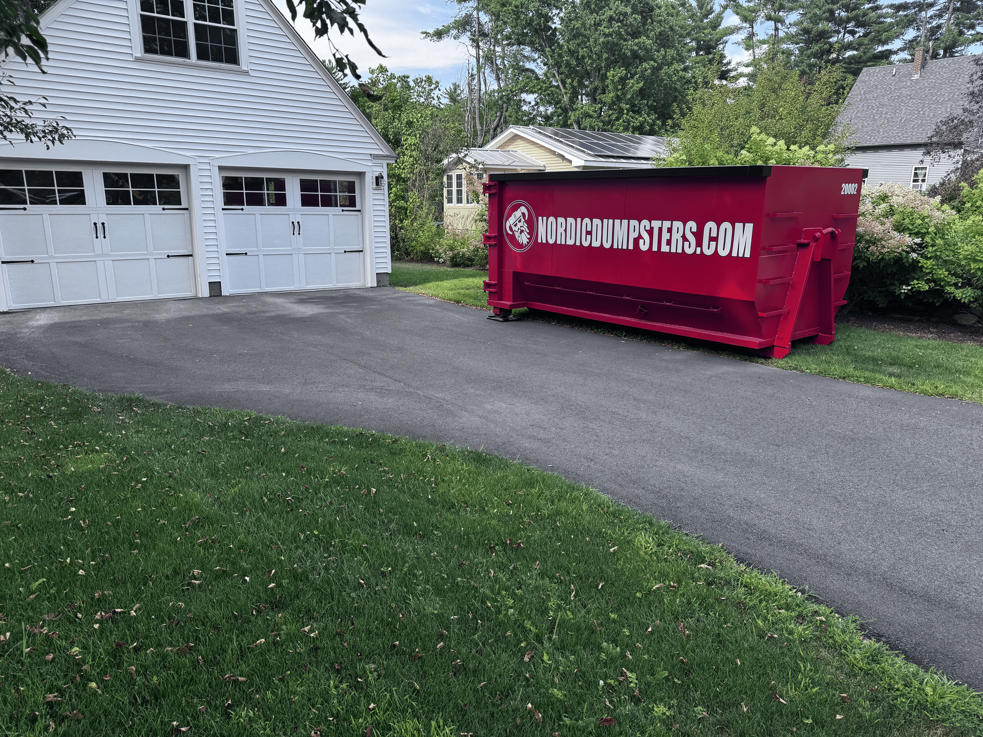 Red trash bin with black lid and red handle, on asphalt pavement near a white truck.