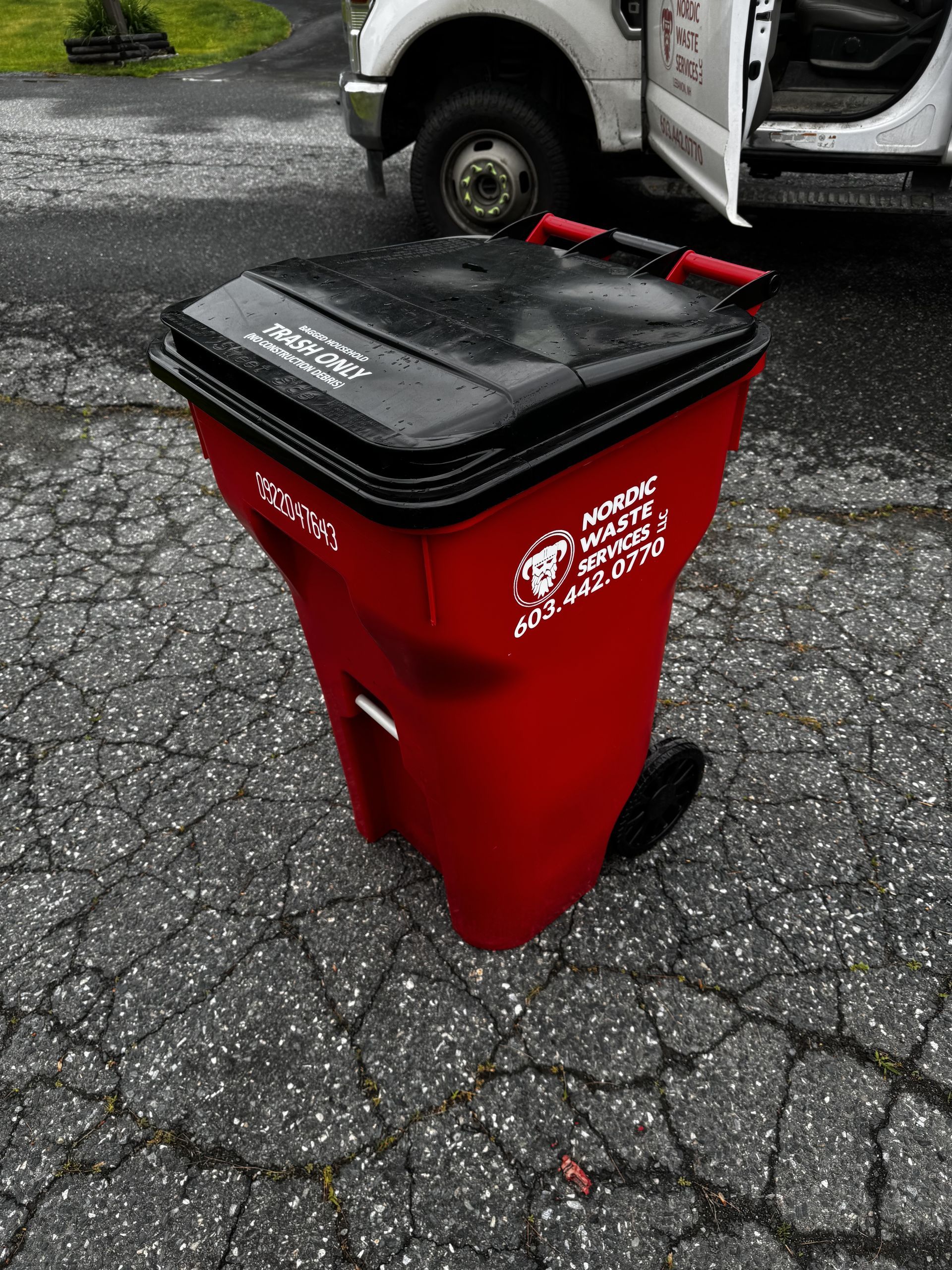 Red trash bin with black lid and red handle, on asphalt pavement near a white truck.
