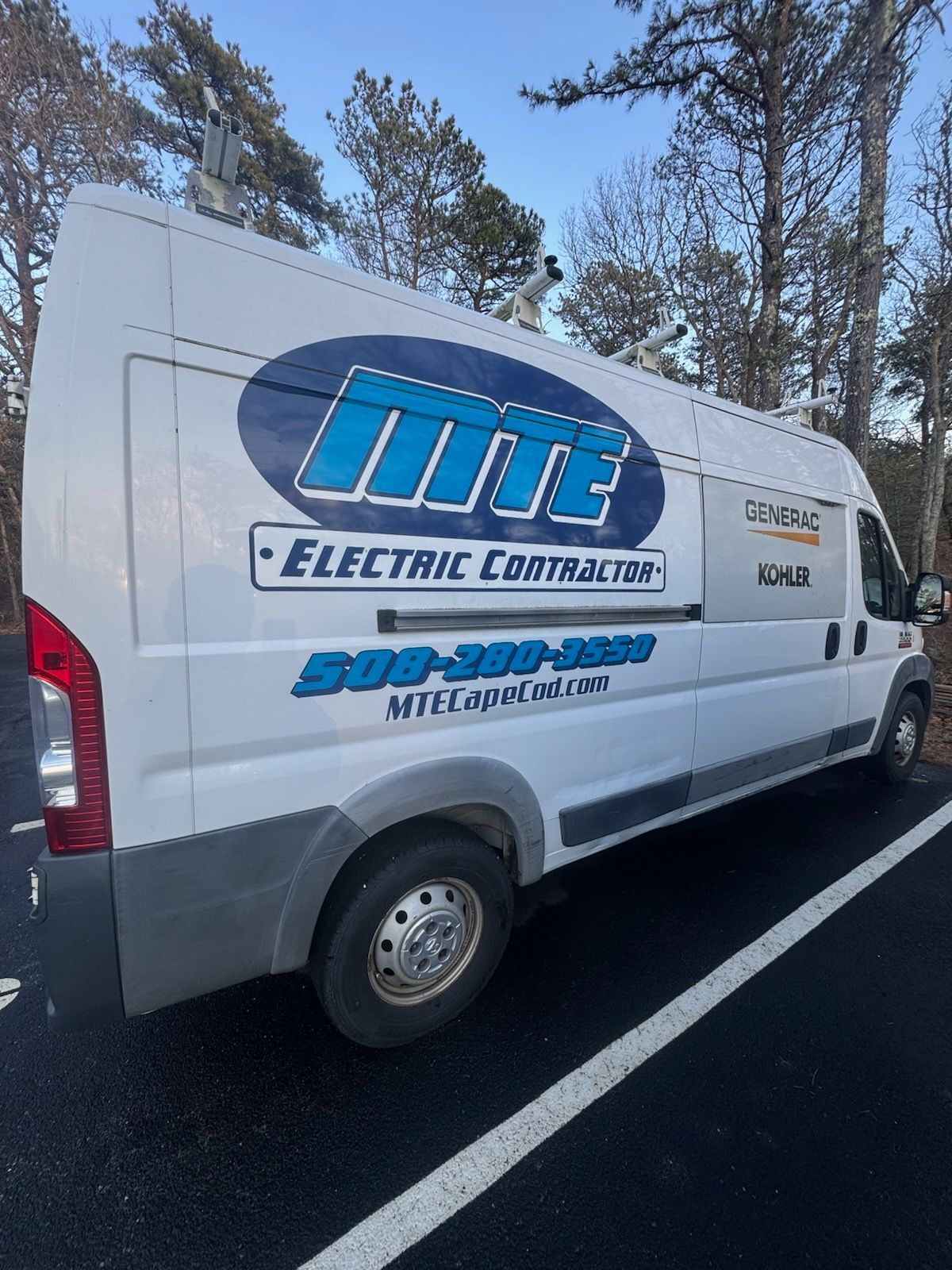 White electric company van with blue logo parked on a road beside trees