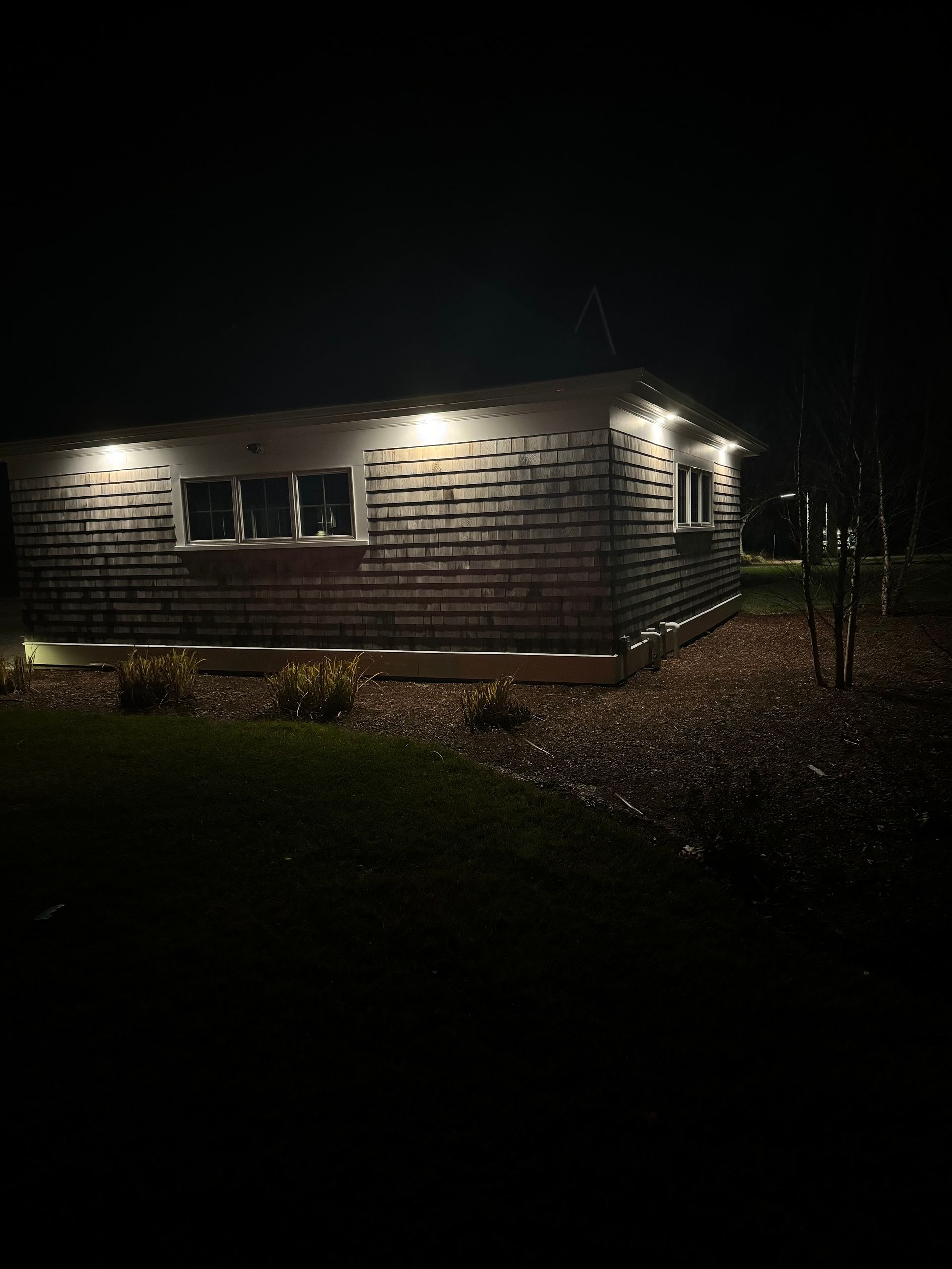 Small house at night, lit by exterior lights, with a dark yard and leaf-covered ground.