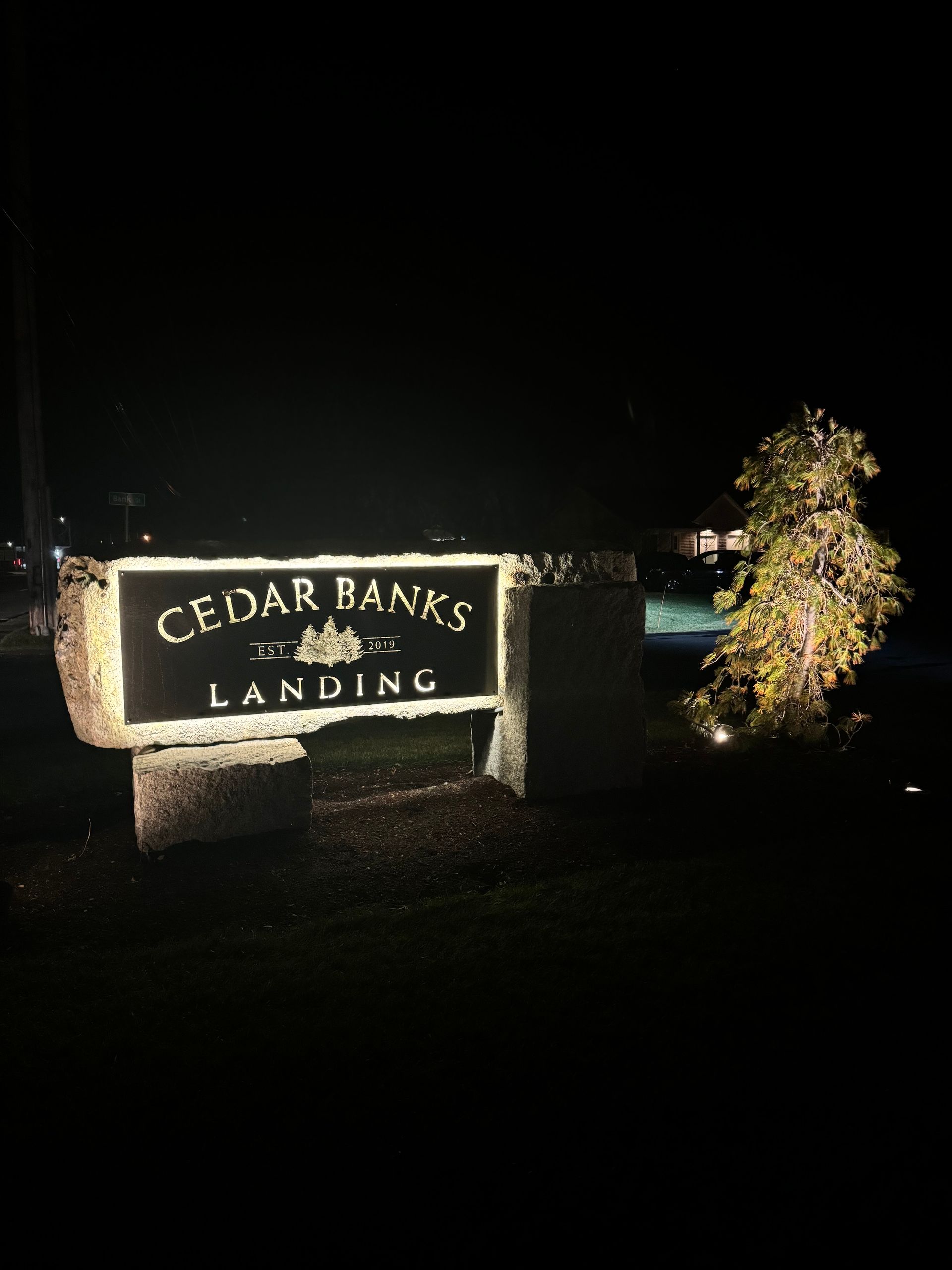 Lit Cedar Banks Landing sign at night with flowers beside it