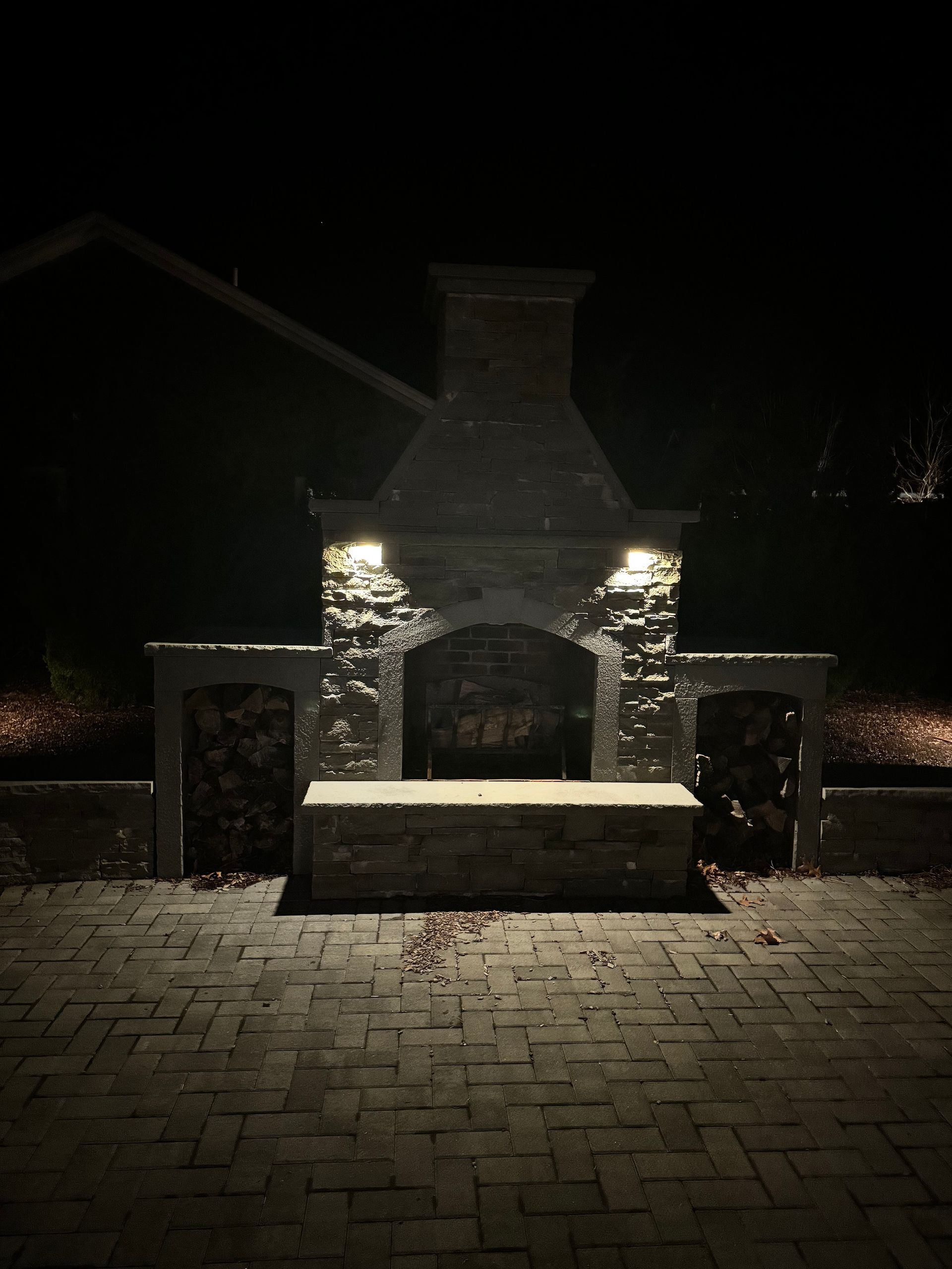 Stone outdoor fireplace with lit sconces and a wooden bench in a dark courtyard.