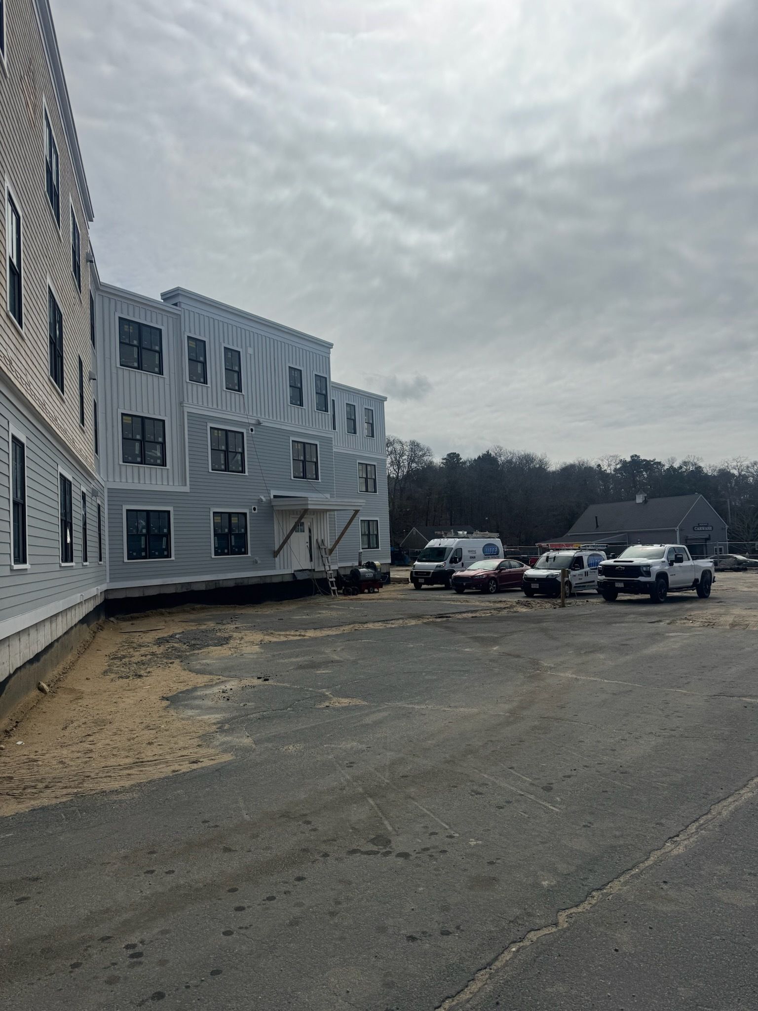 White apartment building beside an empty gravel lot with several parked cars under a cloudy sky
