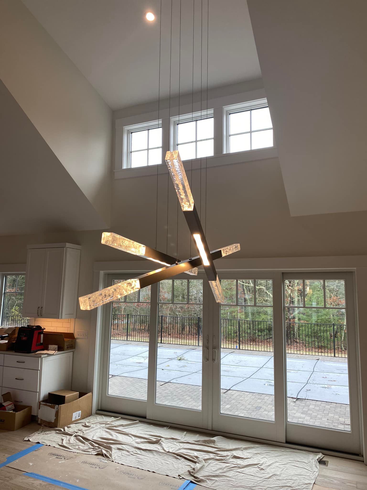 Modern living room with a starburst chandelier, tall windows, and glass doors opening to a snowy backyard.