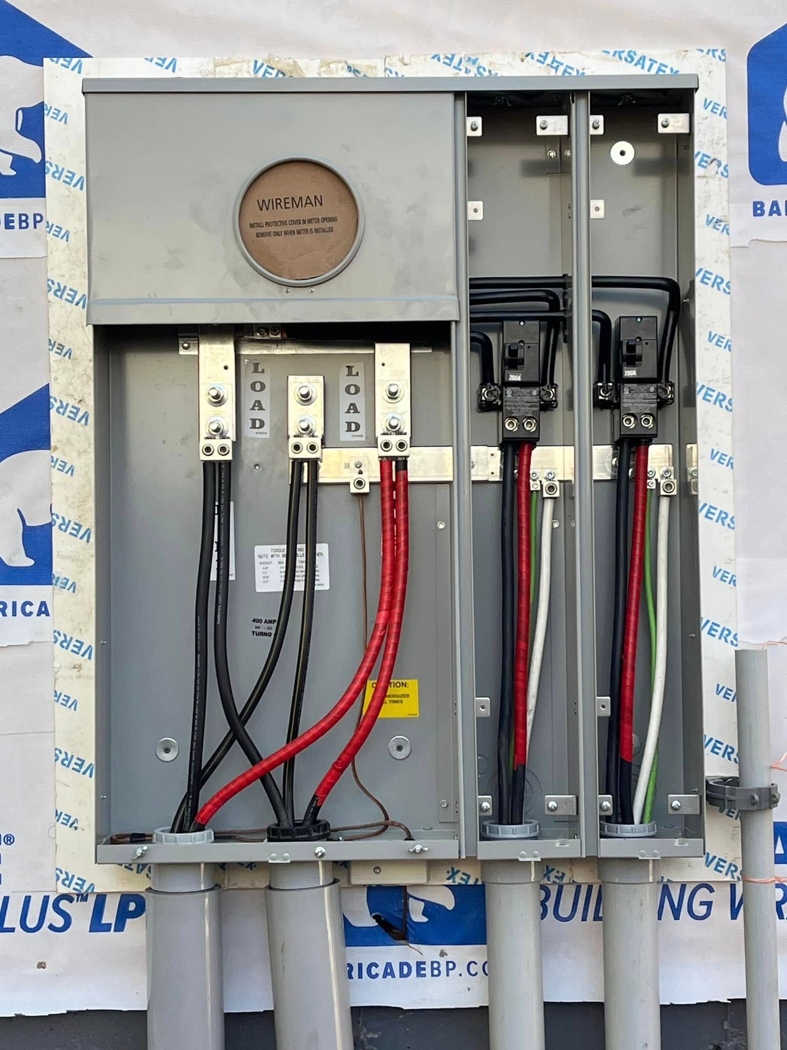 Open electrical service panel with exposed wiring and meters on an exterior wall.