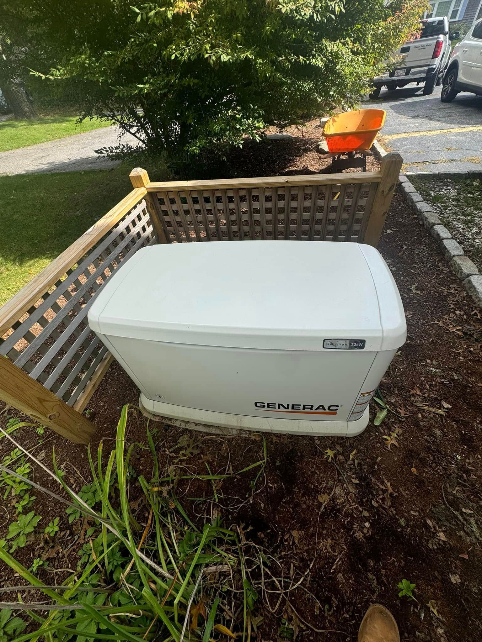 White portable generator behind a wooden barrier on a muddy yard near trees and a driveway.
