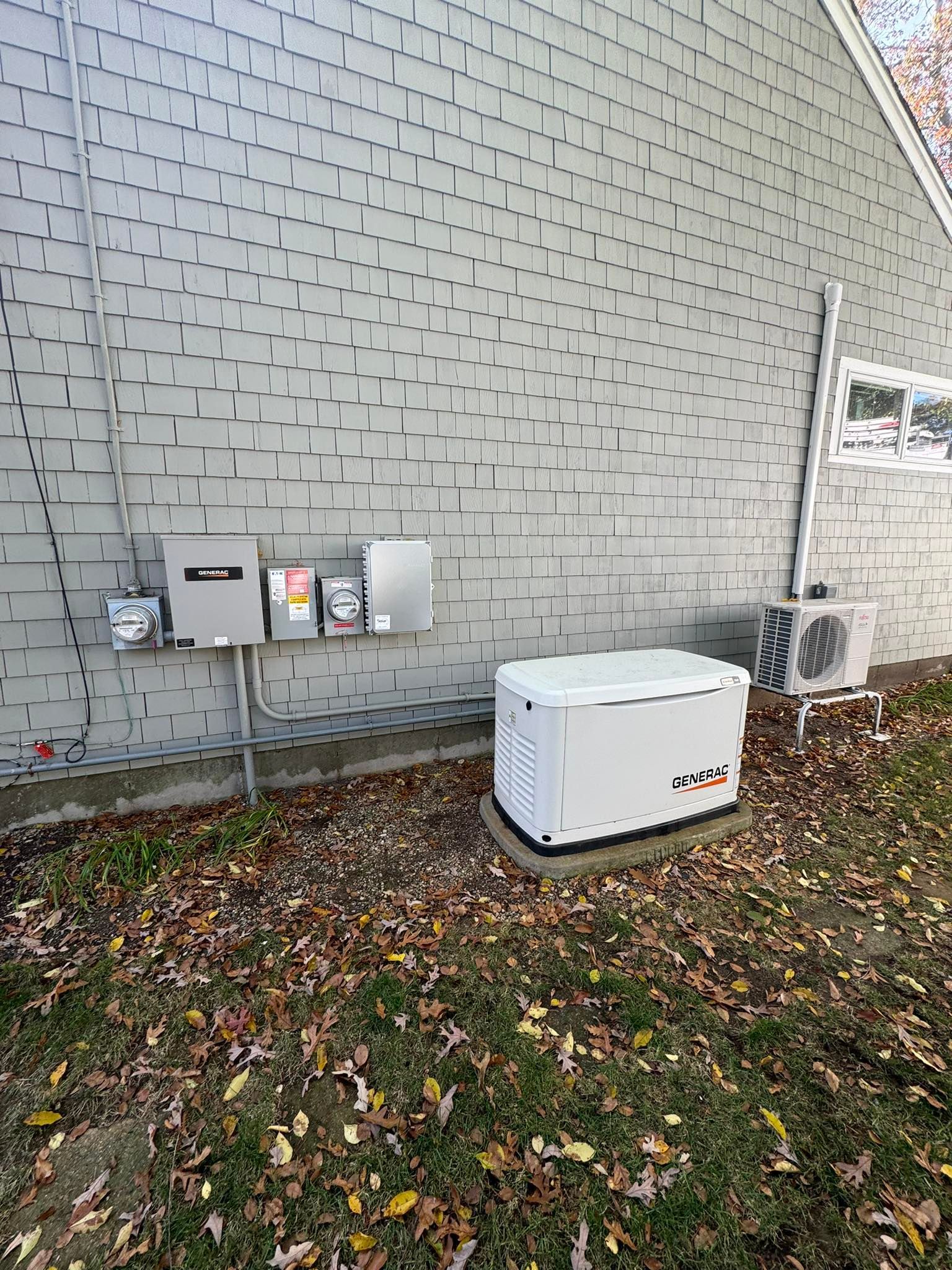Side of a house with utility boxes, an air conditioner, and a white generator on a leaf-covered ground.