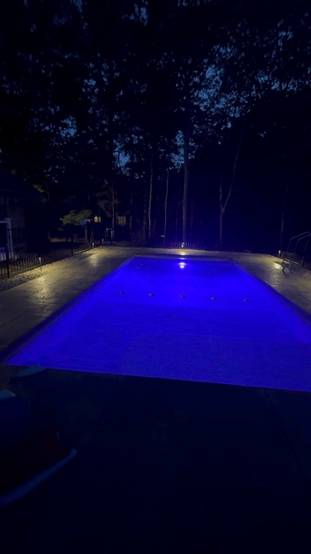 Blue-lit swimming pool at night with surrounding deck and trees in the background
