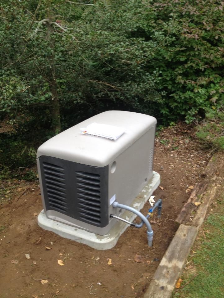 Gray outdoor generator unit on a concrete pad beside a garden path, with hoses attached.
