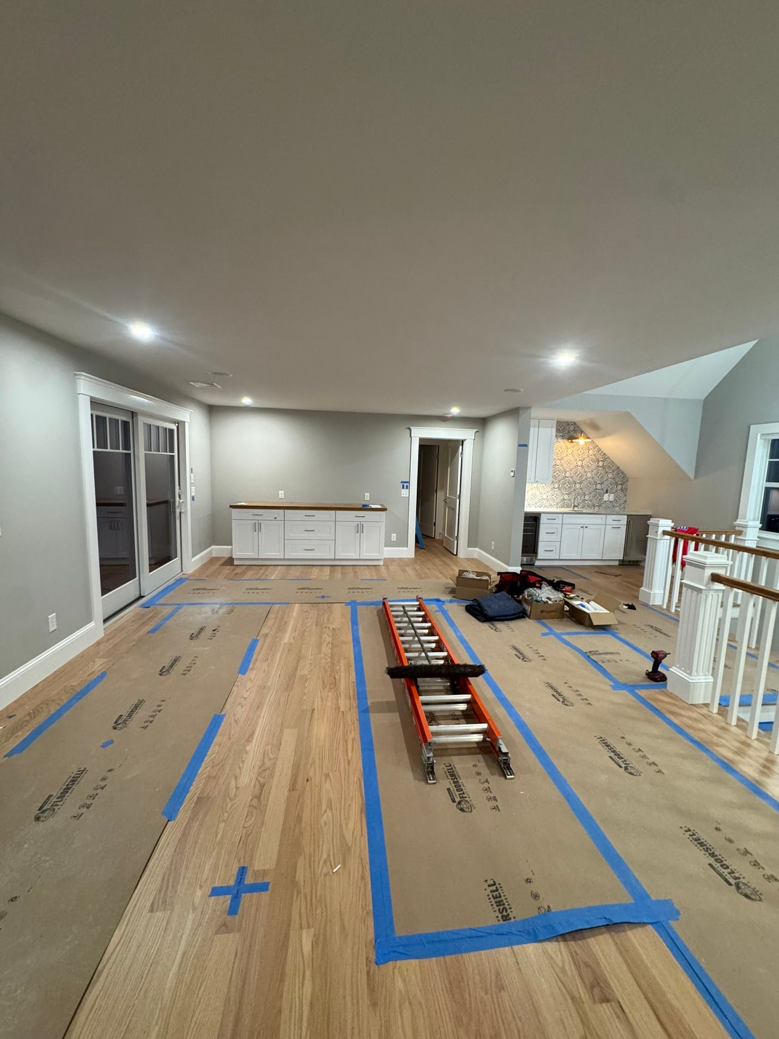 Living room under renovation with flooring taped off, tools, and furniture against the walls.