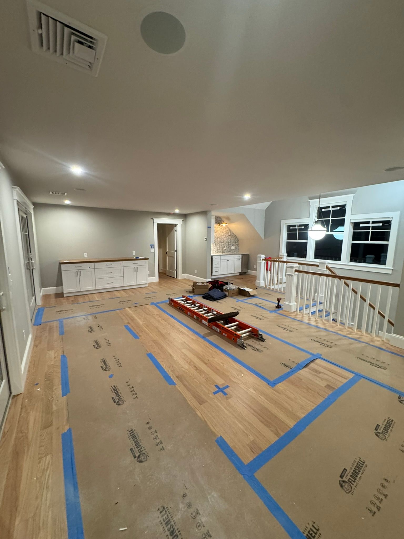 Living room under renovation with taped floor protection, scattered tools, and exposed windows and railing.