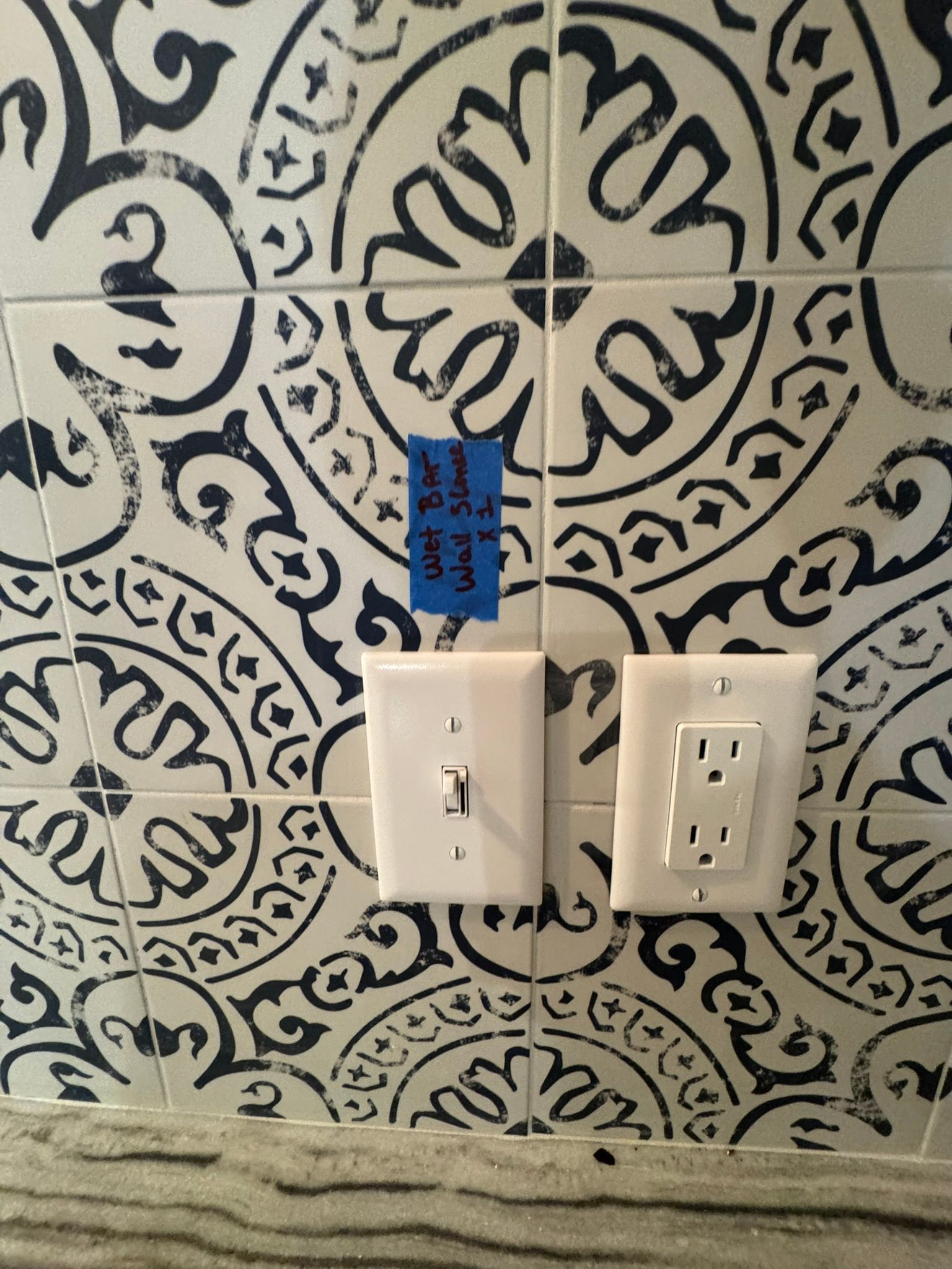 Wall with black-and-cream patterned tile, two white outlets, and a blue tag above one switch￼
