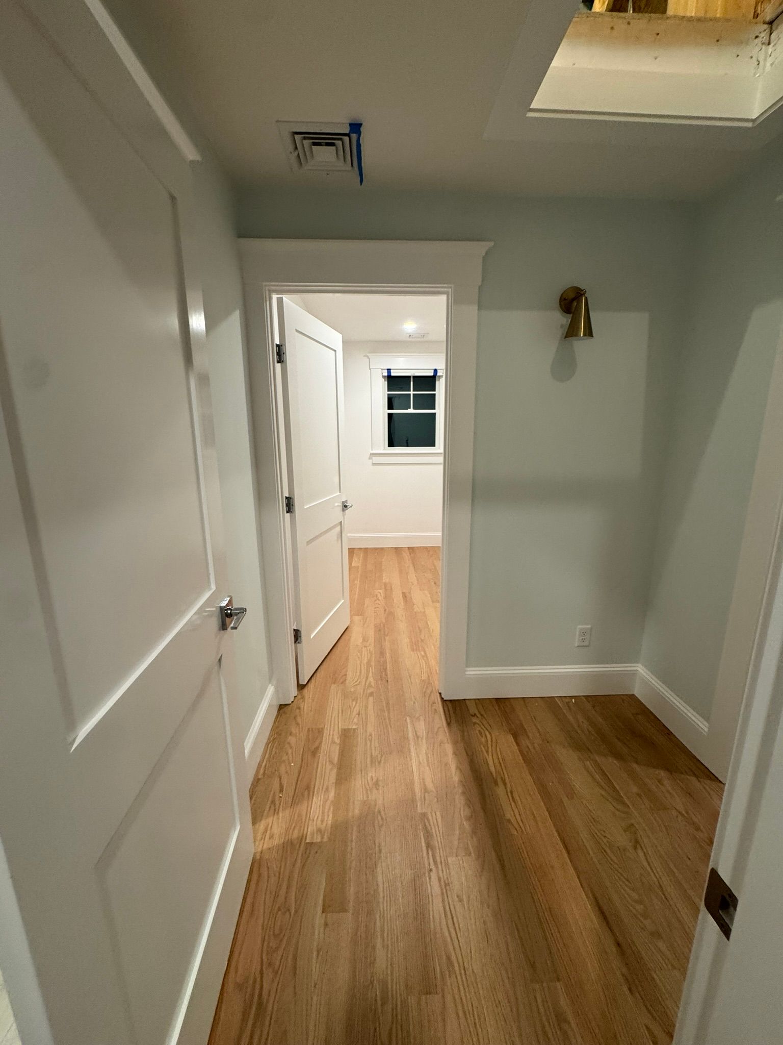 Empty hallway with white doors, light gray walls, and wood flooring leading to a room with a window.