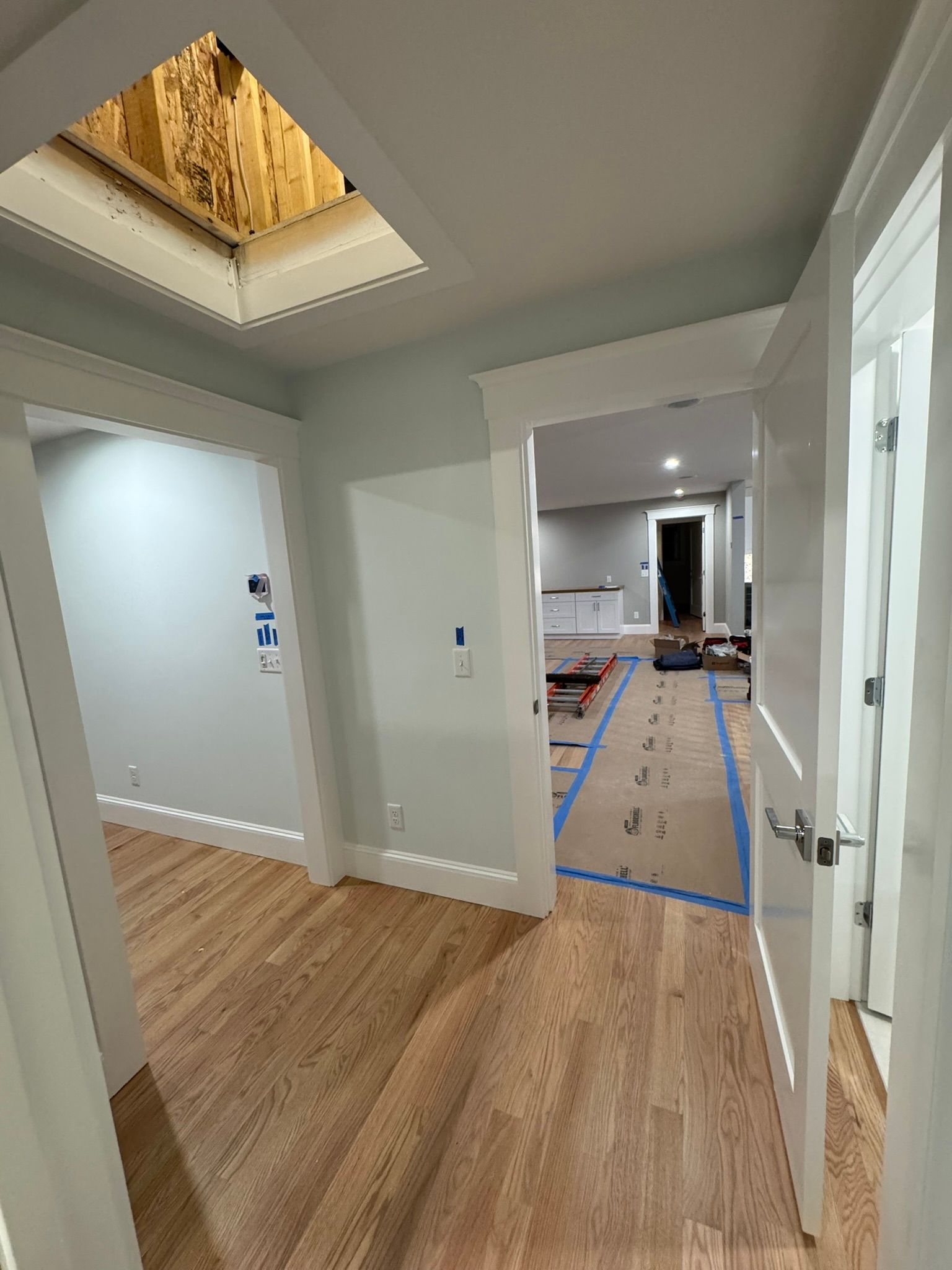 Hallway in a partially renovated home with wood floors, white walls, taped floors, and an open attic hatch.