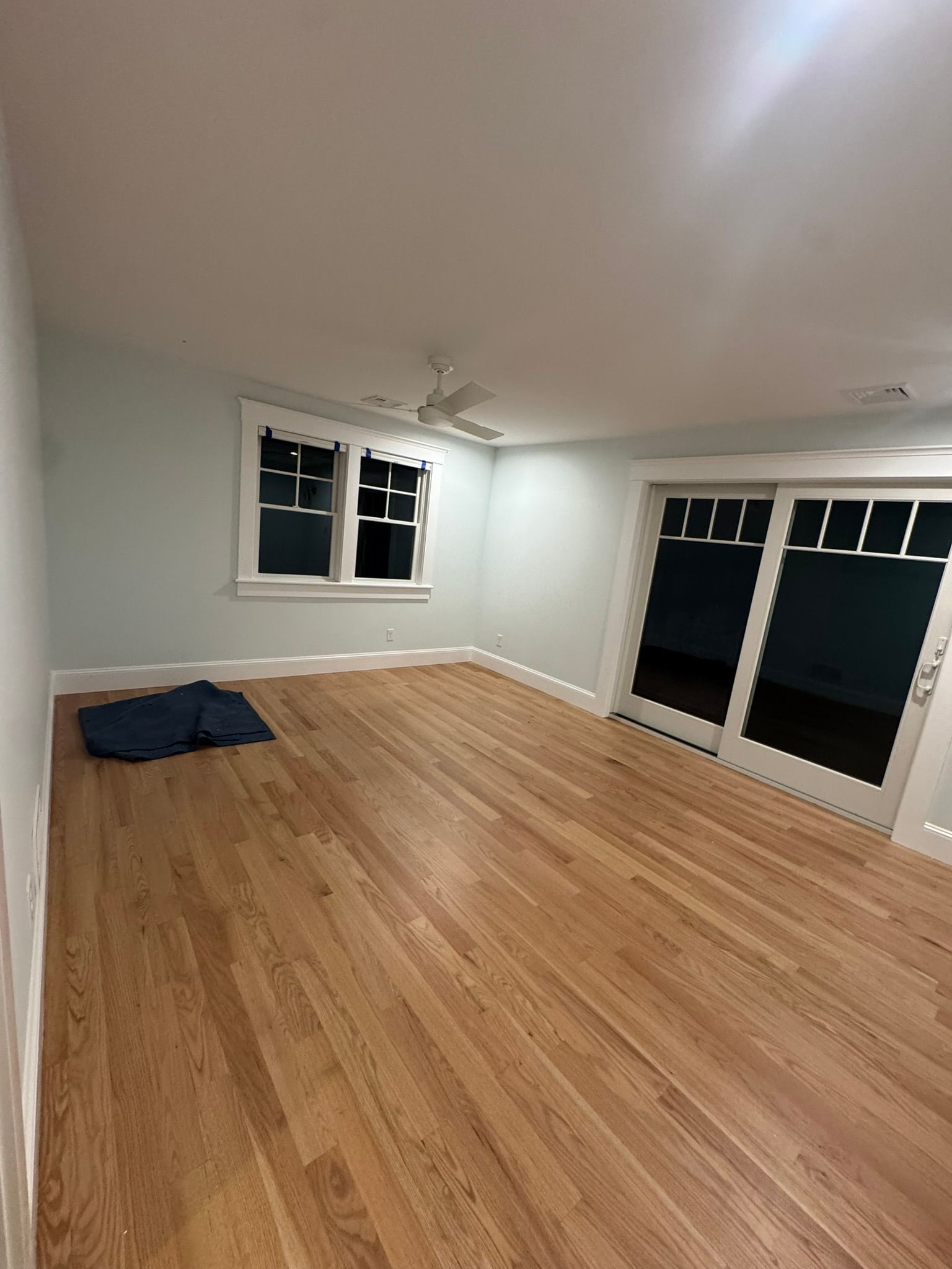 Empty living room with hardwood floors, white walls, windows, and sliding glass doors at night.