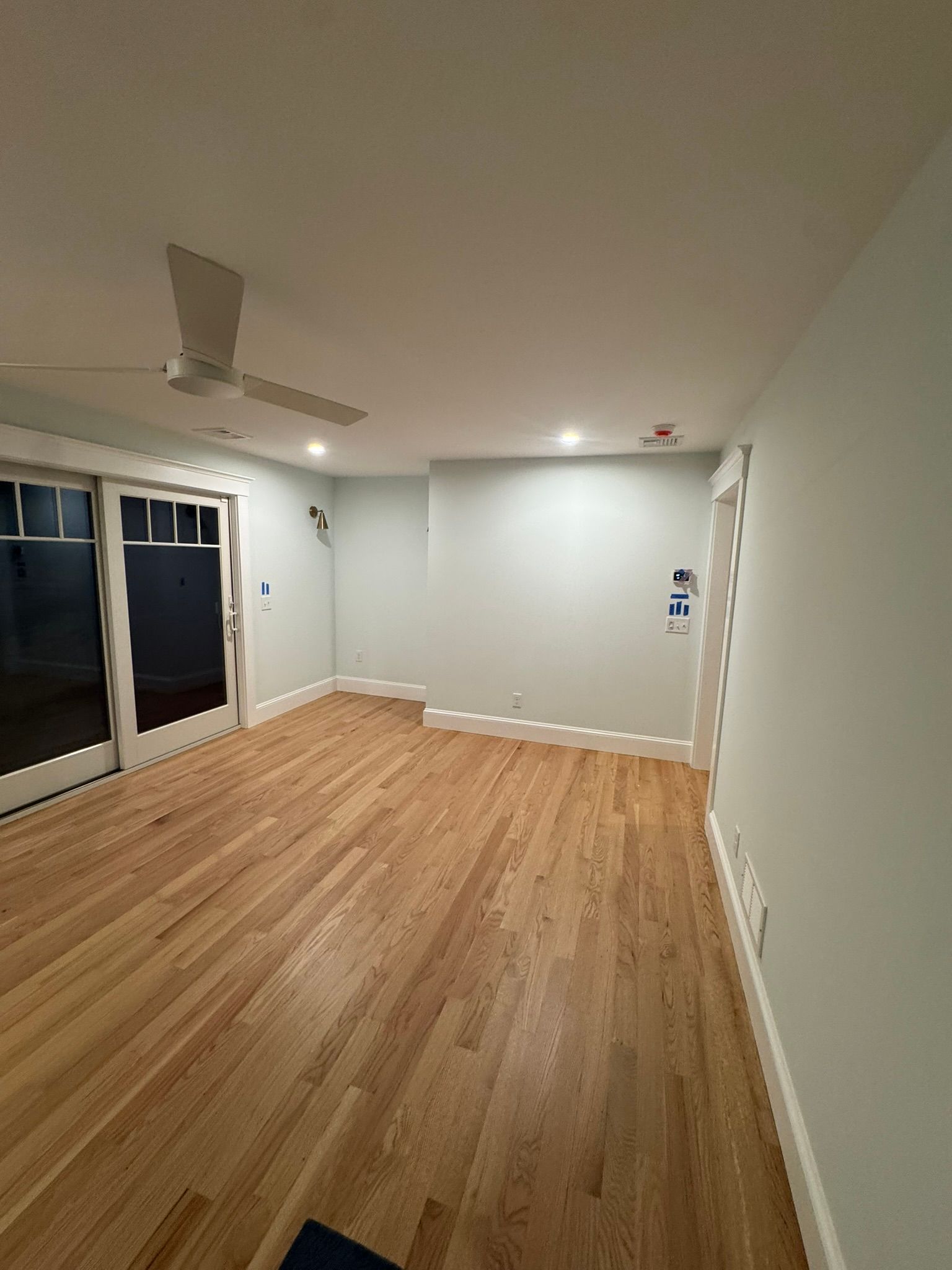 Empty room with light wood floor, white walls, ceiling fan, and sliding glass doors at left.