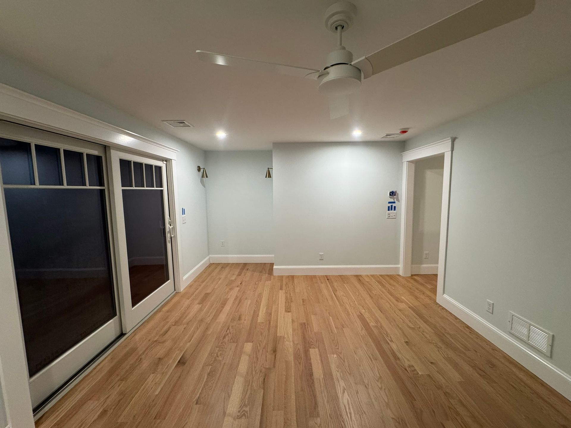 Empty living room with wood floors, light gray walls, ceiling fan, and sliding glass doors.