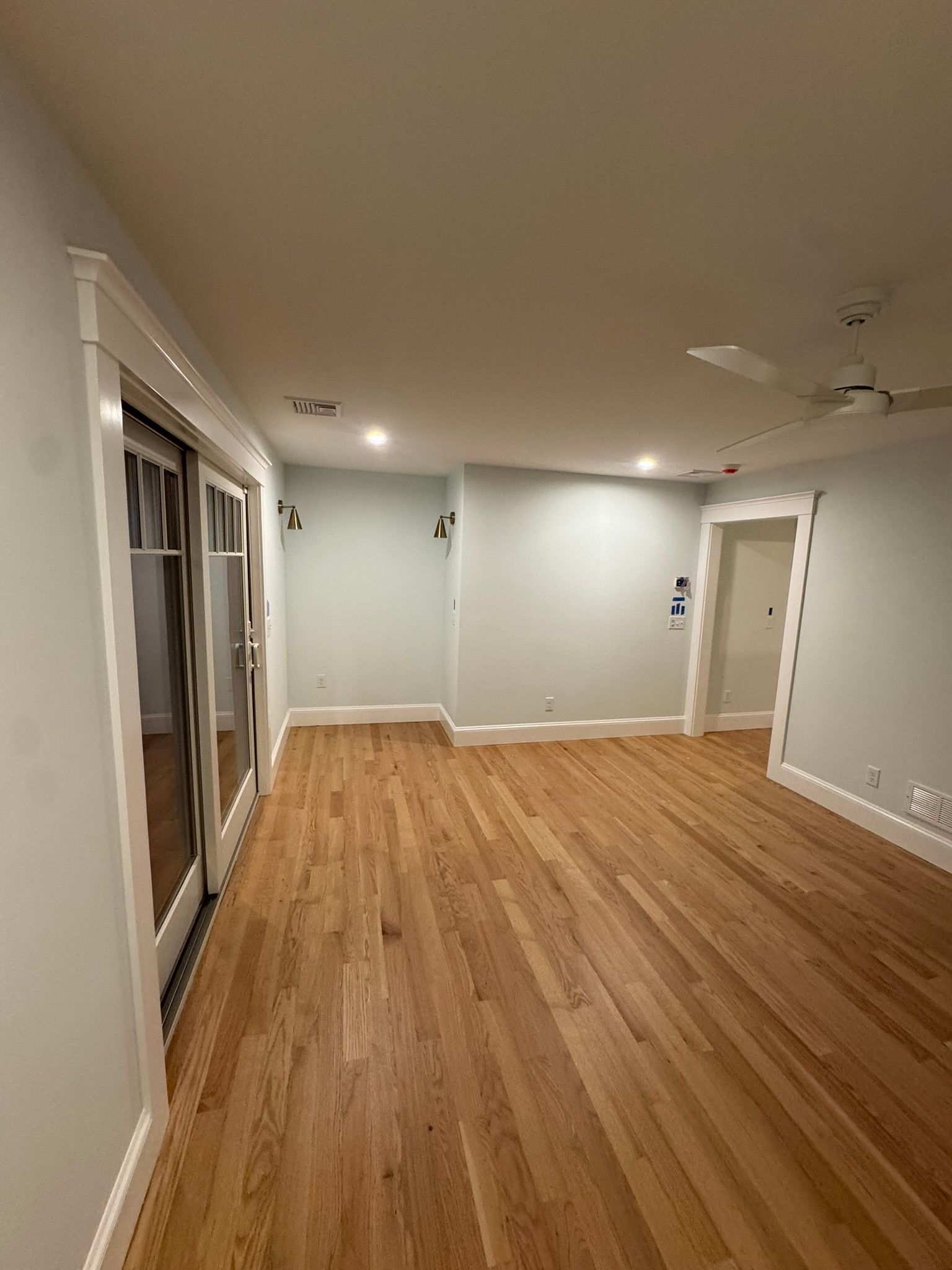 Empty living room with hardwood floors, white walls, sliding glass doors, and a ceiling fan.