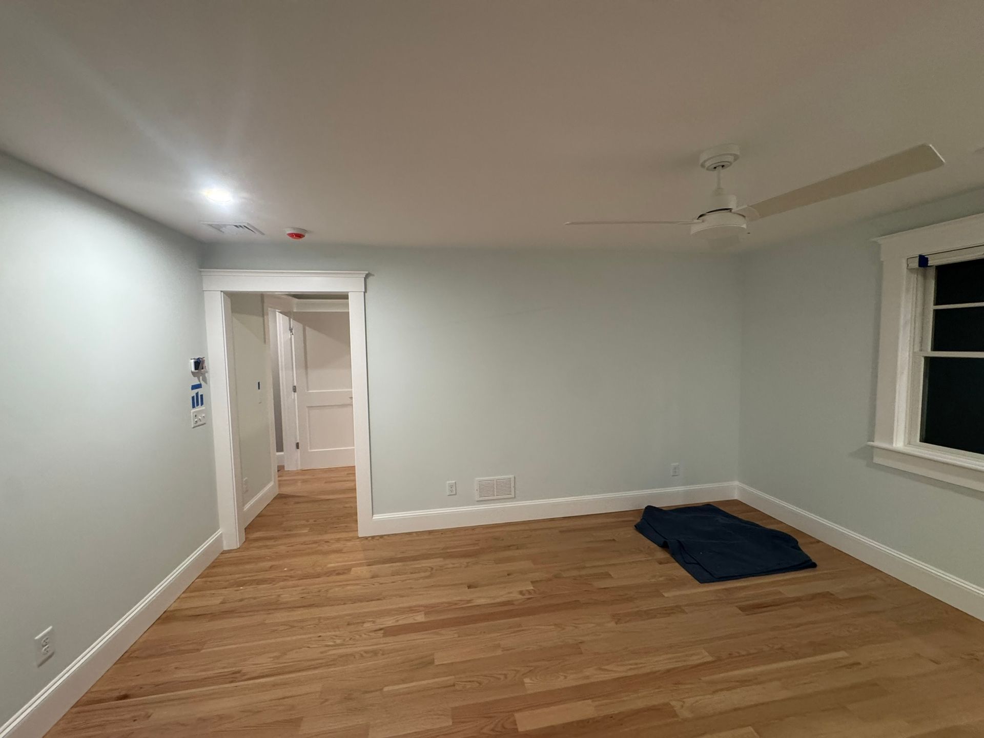 Empty unfurnished room with wood floor, white walls, ceiling fan, and a small blue mat near the window.