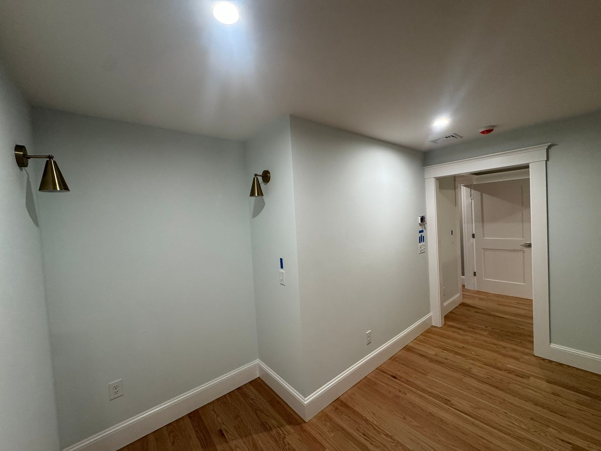 Empty hallway with light gray walls, wood floors, and a doorway to another room.