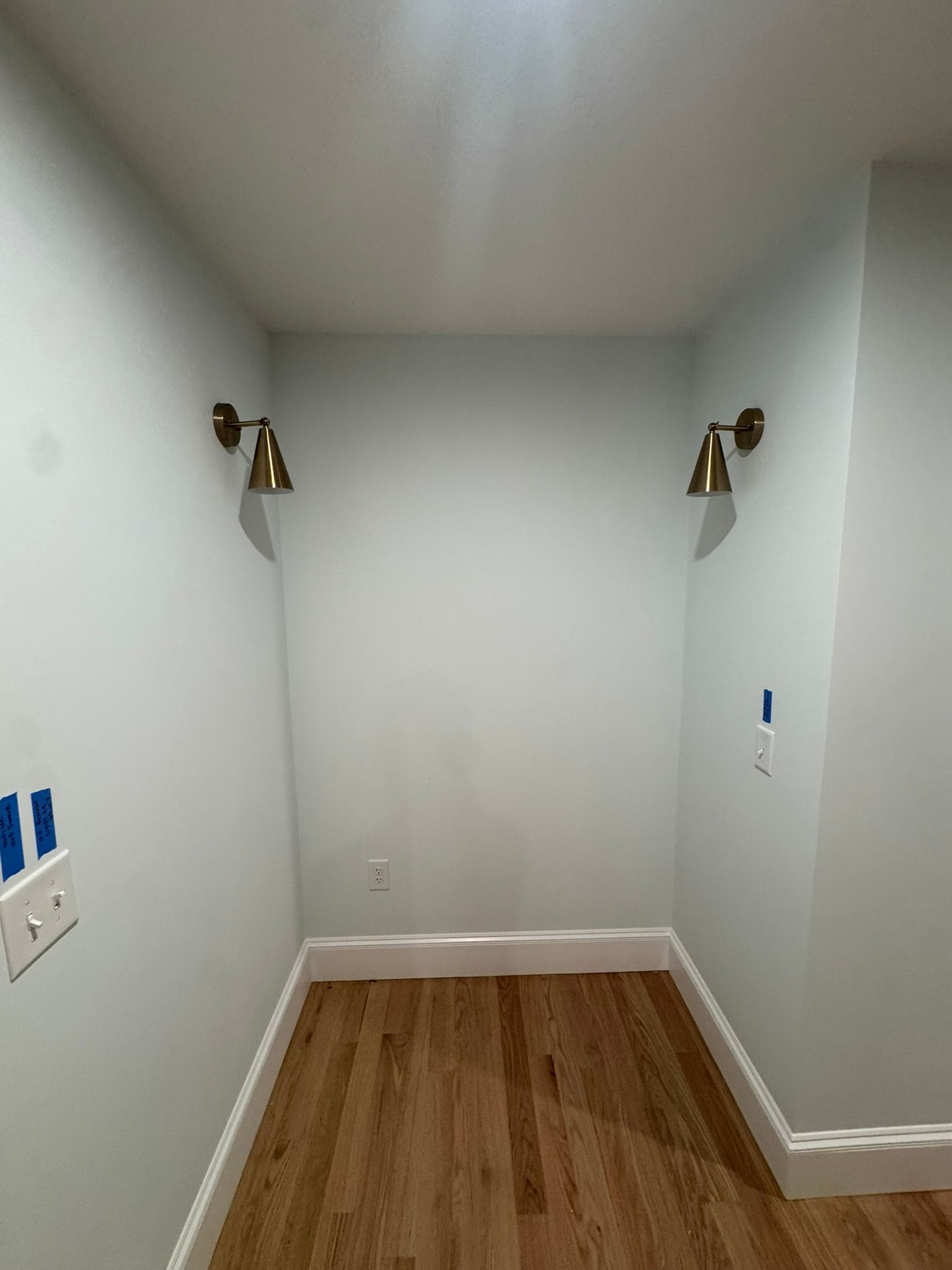 Empty narrow room with white walls, wood floor, and two wall sconces on the back wall.